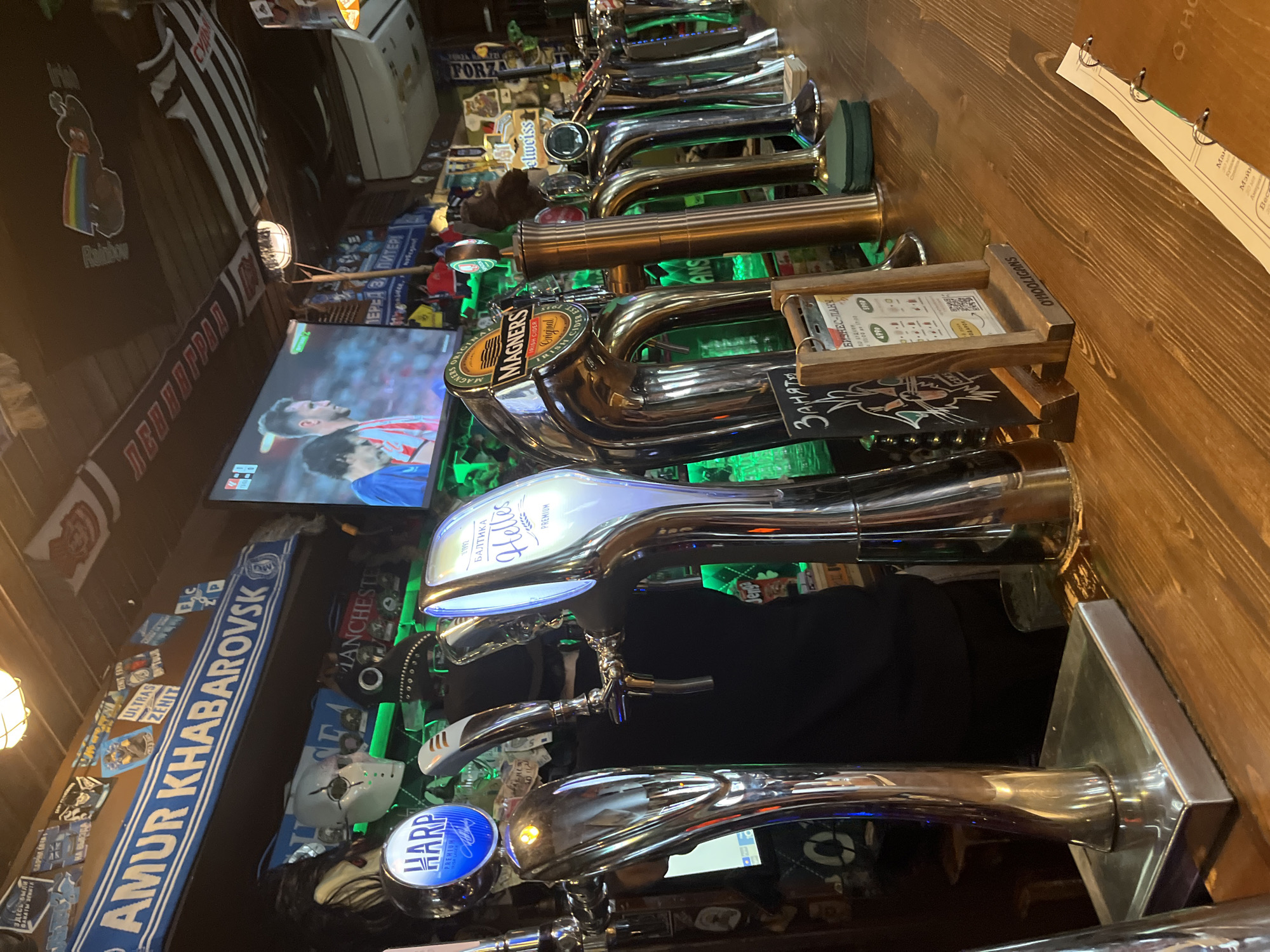 This image depicts the interior of a dimly lit bar or pub, characterized by a long wooden counter featuring a prominent row of shiny chrome beer taps. The lighting is low, with green and blue LED accents illuminating the taps, suggesting an evening or nighttime setting.
Numerous beer taps are visible, bearing brand logos such as "Magners Original Irish Cider," "БАЛТИКА Helles PREMIUM" (Baltika Helles Premium), and "HARP PREMIUM." One tap also has "elweiss" written on it. Behind the taps, a television screen displays a live football (soccer) match, with two players clearly visible.
The walls are richly decorated with various sports memorabilia, including scarves, banners, and patches. Notable texts on these items include "AMUR KHABAROVSK," "ULTRAS ZENIT," and "ФАНАТЫ ЗЕНИТА" (Fans of Zenit), indicating a strong allegiance to Russian sports clubs. Other details include a "MANCHESTER" patch, a "FORZA" banner, a small banner reading "ЗДЕСЬ БЫЛИ" (Here were), and a plush toy with a rainbow design labeled "Rainbow." A white hockey-style mask is also part of the decor.
On the counter, there are small wooden menu holders. One clearly displays the name "O'HOOLIGANS," and another sign reads "БИЗНЕС-ЛАНЧ 07:00 4990" (Business Lunch 07:00 4990) and "Занято" (Occupied), indicating a reserved space. A hanging menu shows sections like "Ман," "Мэйн," "30 мя," and "Becz." The prevalence of Russian text and references to Zenit FC, a club from Saint Petersburg, Russia, strongly aligns with the provided location context. The scene suggests a typical pub atmosphere for watching sports and socializing.
