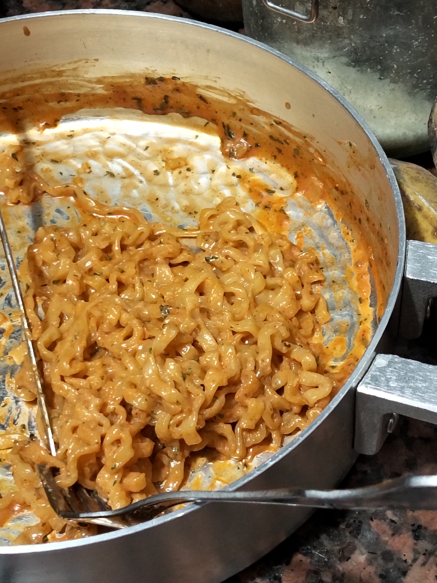 This is a close-up, eye-level shot of a pot filled with cheesy noodles, likely ramen or instant noodles, coated in a creamy orange sauce. Two forks are visible, one partially submerged in the noodles and the other resting on the rim of the pot. The pot itself is silver with two handles. The background is blurred but appears to include a metallic container and possibly some fruit, such as bananas. The setting is a kitchen counter, indicated by the speckled granite surface. The image quality is good, with clear focus on the food and utensils. There is no indication of the specific location within Davao City, Philippines.