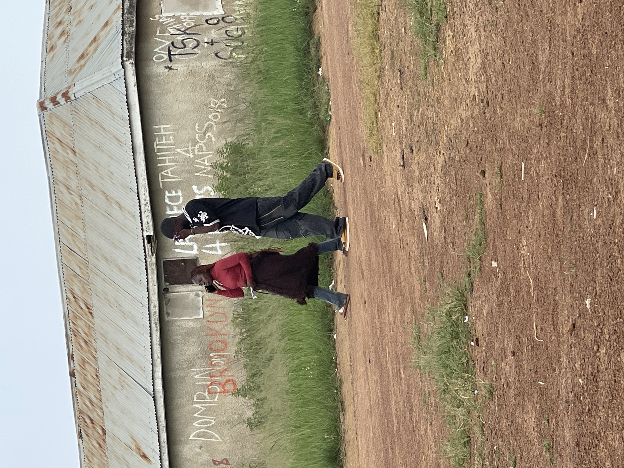 In the city of Durumi, Nigeria, two individuals walk along a dirt road in front of a weathered, corrugated metal-roofed building. The building's concrete walls are covered in graffiti, with visible words including "DOMBIN," "BROOKLYN," "ECE TAHITEH," and "NAPSS." A woman in a red top and dark skirt is on the left, speaking on a mobile phone. To her right, a man in a black shirt with white lettering and dark pants is also holding a phone to his ear. Tall, green grass grows along the side of the building and the road. The ground is composed of reddish-brown dirt with small stones and sparse vegetation. The sky is overcast.