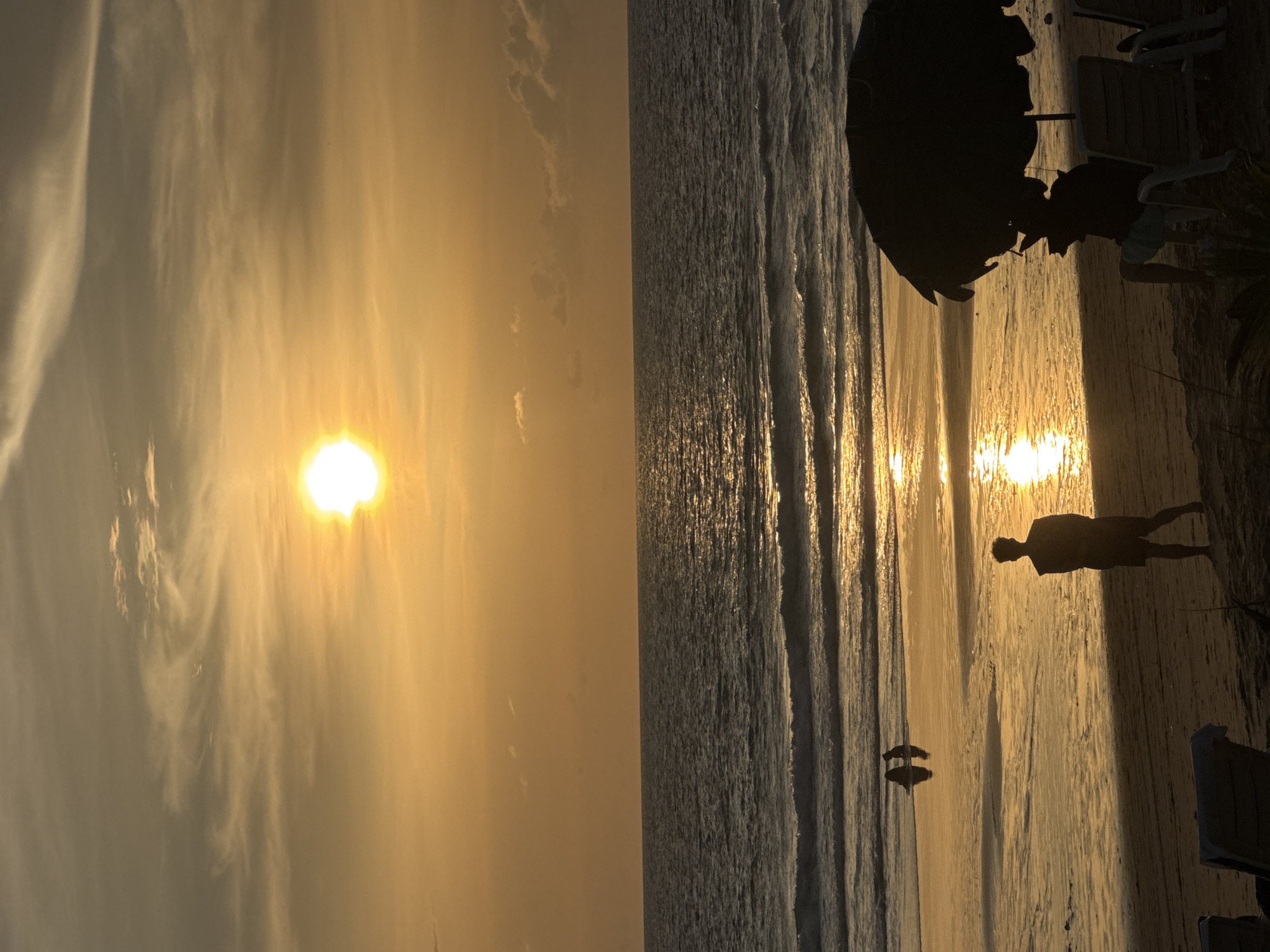 At sunset on Karon Beach, Thailand, the sun is a bright disc low in the sky, casting a warm golden glow across the scene. Wispy clouds streak the sky, catching the light. The ocean stretches to the horizon, with gentle waves lapping the shore, reflecting the sun's brilliance. On the wet sand closer to the water, two silhouetted figures stand side-by-side, their reflections shimmering in the shallows. Further up the beach, a lone silhouetted figure stands facing the ocean. In the foreground, a large dark beach umbrella shades two empty lounge chairs, and another silhouetted figure sits in one of the chairs, looking out towards the sea.