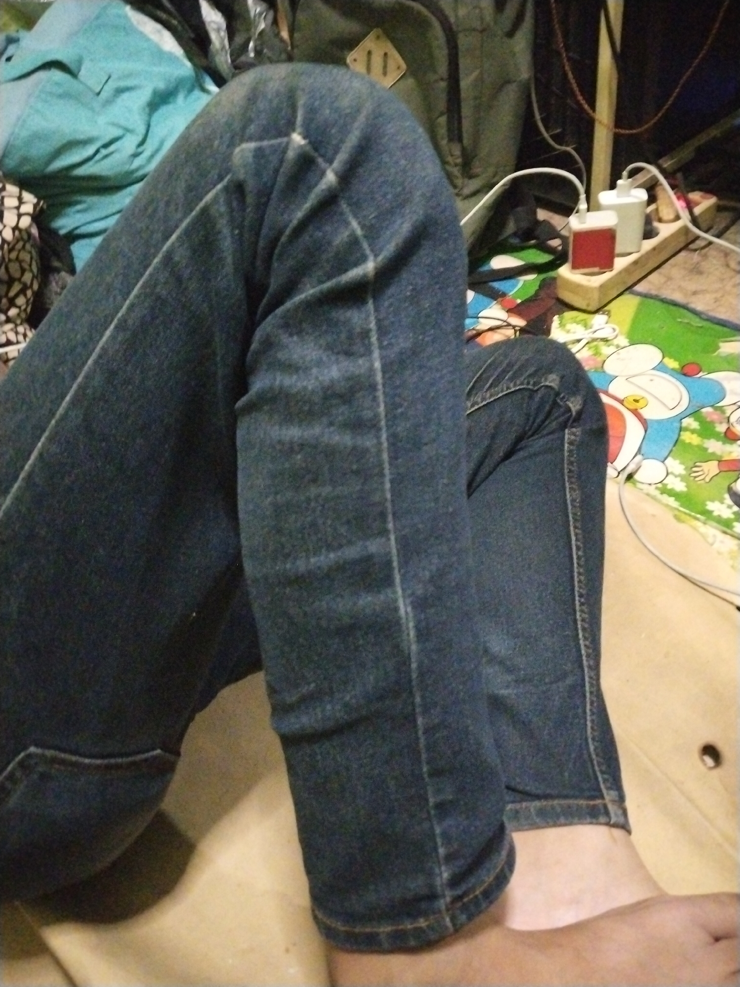 The image shows a close-up view of a person's legs wearing blue denim jeans. The person is sitting or kneeling on a surface that appears to be a light-colored mat or floor. In the background, there are various objects including a teal-colored shirt or fabric, a dark backpack, and a power strip with two charging adapters plugged into it. One adapter is white, and the other is red. White cables are connected to the adapters. A colorful mat with cartoon characters, possibly Doraemon, is visible beneath the power strip and extending towards the person's legs. The scene appears to be indoors, possibly in a dimly lit room, as suggested by the lighting and the presence of electrical outlets and chargers. The focus is on the person's legs and the surrounding objects, with no discernible activity or event taking place beyond the person being in a resting or stationary position. There is no visible text in the image.