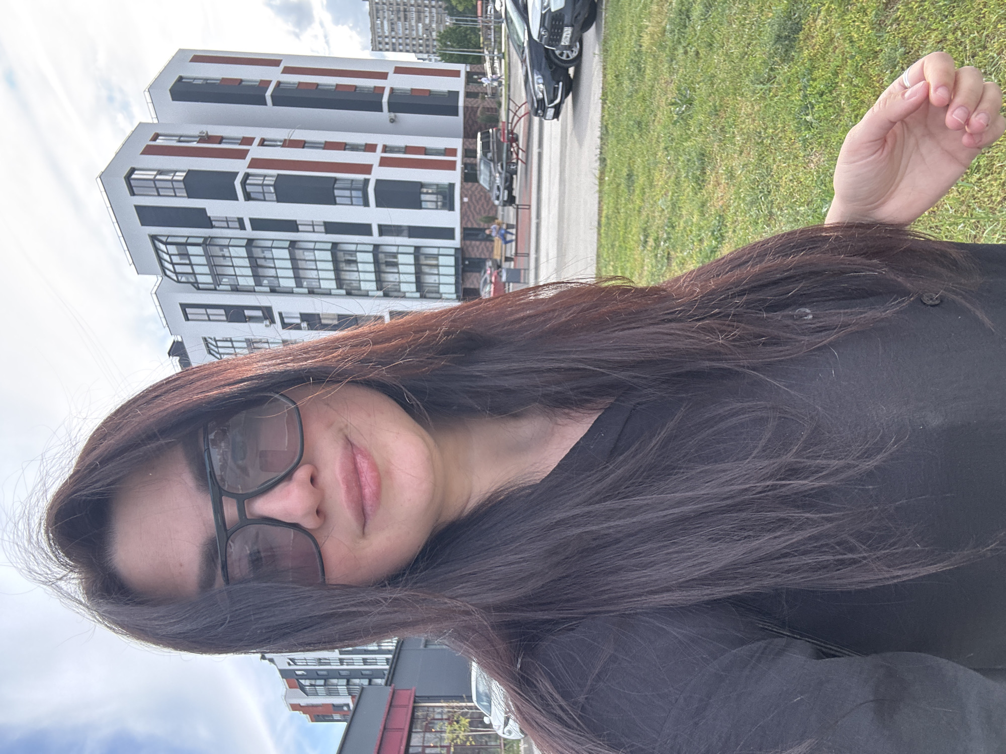 The image shows a woman with long, dark brown hair wearing dark sunglasses and a black shirt. She appears to be taking a selfie. In the background, there is a multi-story building with white and dark grey sections, accented with reddish-brown panels. A street runs beside the building with parked cars. A patch of green grass is also visible. The location appears to be in a city.
