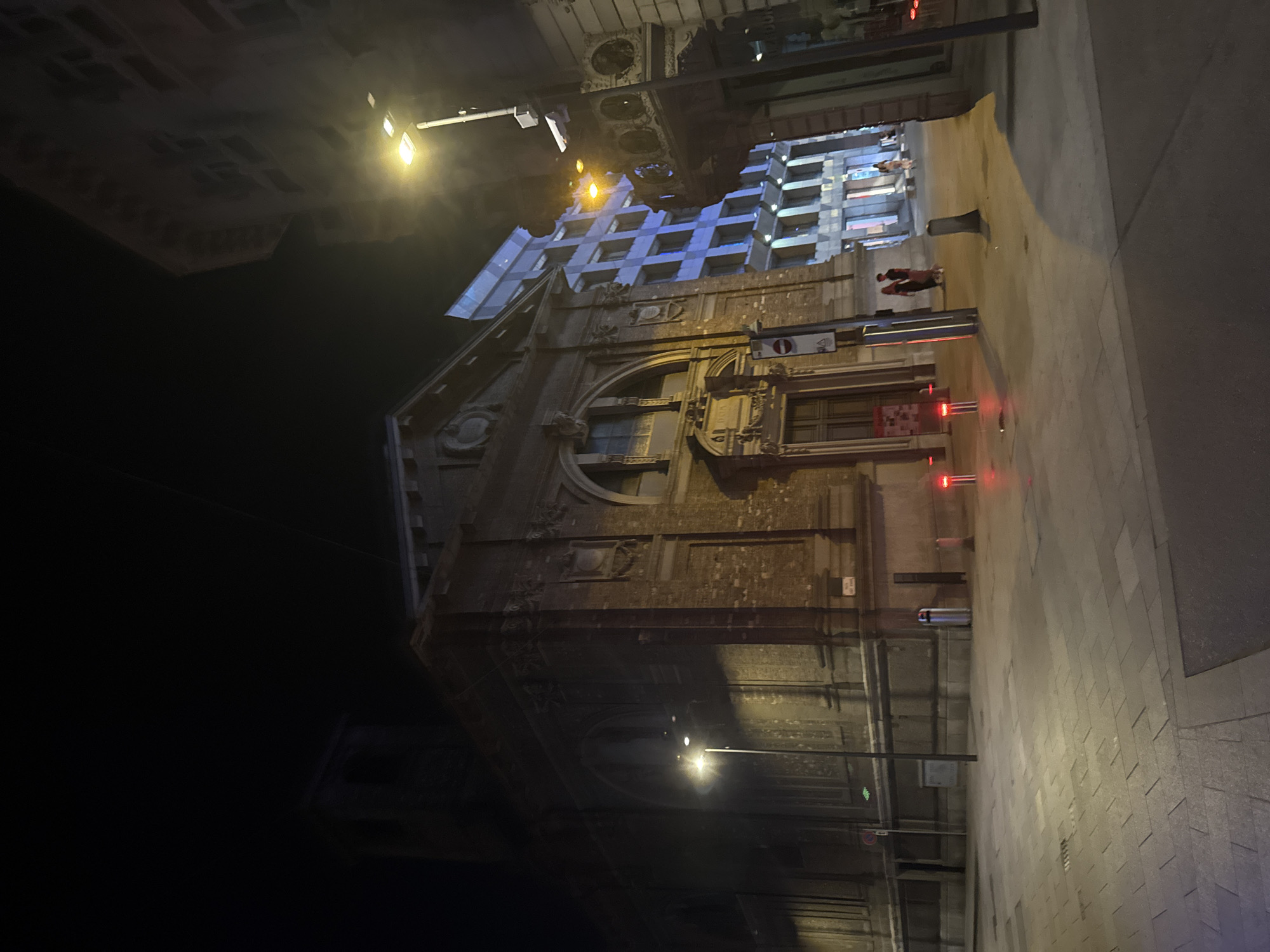The image shows a street scene at night in Lugano, Switzerland. A large, ornate building with a dark brick facade and arched windows is prominent on the left. To its right, a modern building with blue-lit windows is visible. A street lamp with a bright light illuminates the scene. The street is paved with rectangular stones, and there are several bollards and a trash can visible. In the distance, a few people are walking on the sidewalk. The overall atmosphere is dimly lit and somewhat deserted, suggesting late evening or early morning.