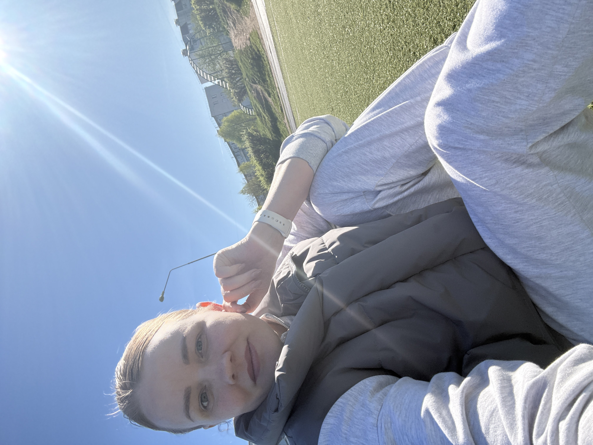 The image captures a fair-skinned woman with blonde hair and a white wristwatch, seated outdoors in a city. She is wearing a grey hooded sweatshirt and a grey vest. She is holding something near her ear. The background shows a green surface resembling a field, along with a building, trees, a fence, and a clear blue sky with the sun shining brightly. The image appears to have been taken in the city of Chaikovsky, Russia.