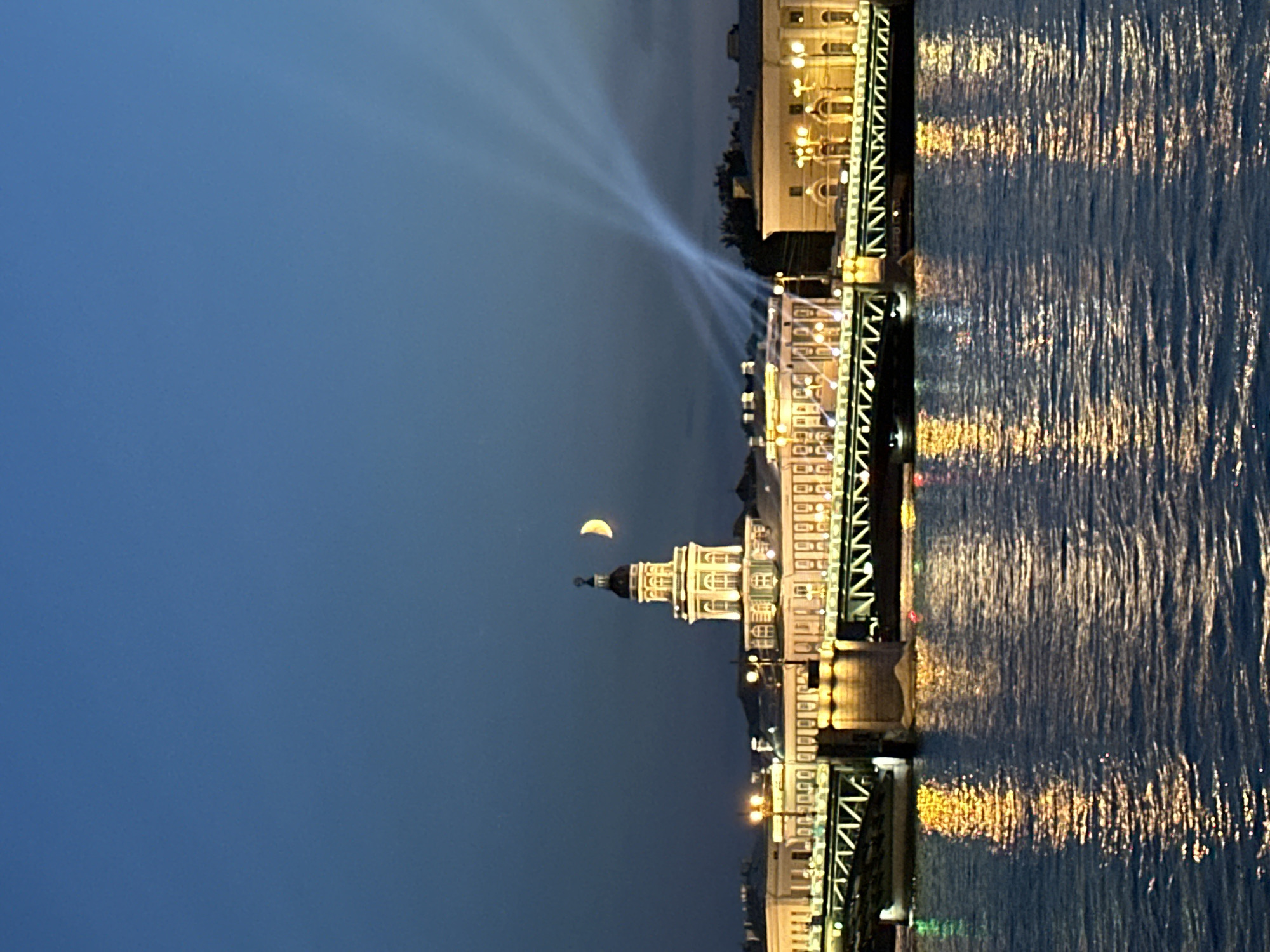 The image depicts a nighttime view of the Neva River in Saint Petersburg, Russia, featuring the Palace Bridge (Dvortsovy Most). Illuminated buildings line the riverbank, including the Kunstkamera with its distinct tower, visible in the center. A crescent moon hangs in the deep blue sky above the Kunstkamera. Searchlights project beams of light into the sky from the right side of the frame. The bridge itself is lit, with its intricate metalwork clearly visible. The river reflects the lights of the city, creating shimmering patterns on the water's surface. There are no people or discernible actions visible in the scene. The overall atmosphere is serene and picturesque.