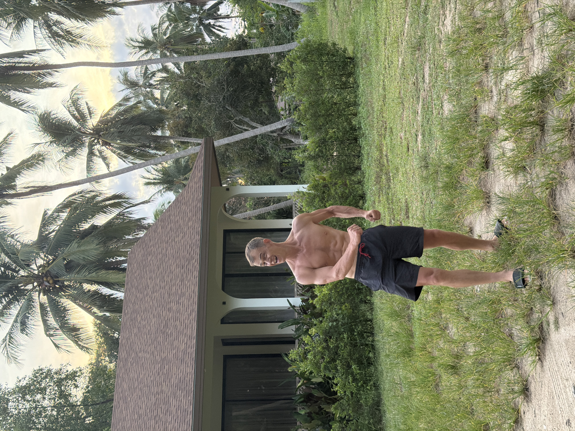 A shirtless man with short blond hair and a tan is posing with his arms bent and fists clenched, looking to his left with an open-mouthed expression of excitement. He is wearing black swim shorts with a red drawstring and dark sandals. He stands in a grassy yard in front of a white house with a brown roof and large windows. Palm trees are visible in the background against a pale sky with hints of yellow and orange, suggesting it is either sunrise or sunset. The environment appears tropical, consistent with the location of ตำบลเกาะพะงัน, Thailand. Lush green bushes and trees surround the house and yard.