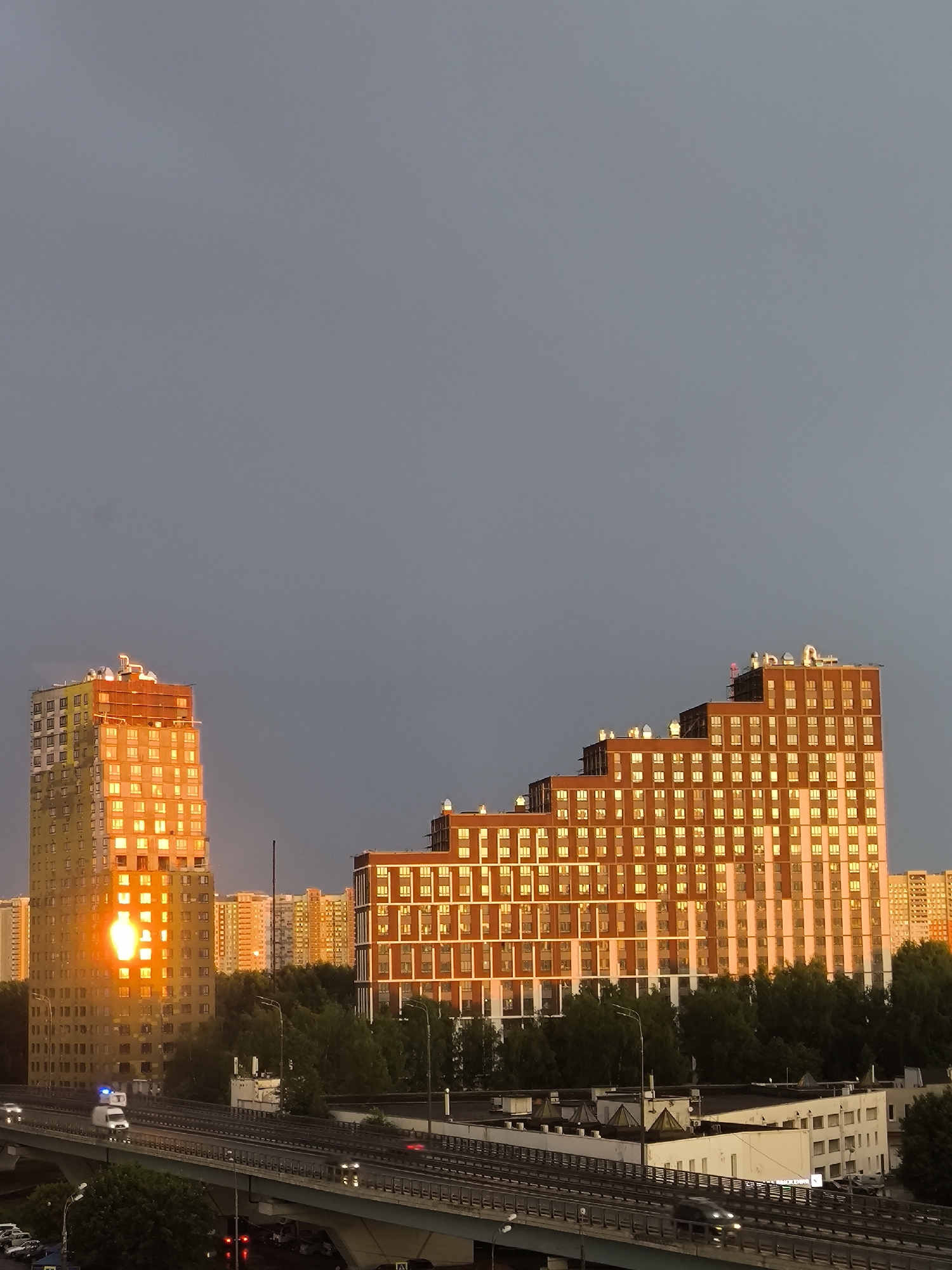 A wide shot captures a city skyline at dusk, dominated by a large, multi-story building with a stepped architectural design on the right, bathed in the warm glow of the setting sun. This building features a repetitive pattern of windows and is constructed with brick and white trim. To the left, a taller, cylindrical building reflects the sun's intense orange light across a significant portion of its facade. In the background, several other residential buildings are visible, also catching the golden hour light. A dense line of green trees separates the buildings from a multi-level highway in the foreground. Cars and a white van with blue flashing lights are driving on the highway. The sky above is a muted gray, indicating overcast conditions or the transition from day to night. The overall scene suggests an urban environment during sunset.