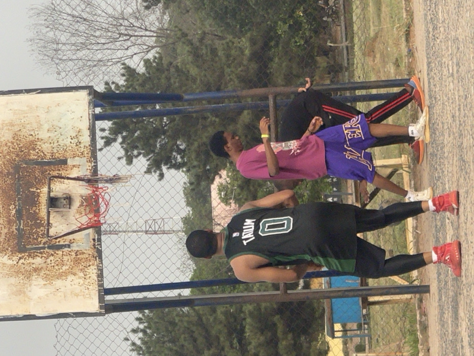 The image depicts an outdoor basketball court in Umunekwu, Nigeria, featuring three individuals and a basketball setup. A heavily rusted, white-colored rectangular basketball backboard with blue support poles stands centrally, fitted with a hoop displaying an incomplete net composed of red, white, and blue segments.
In the foreground, a person is seen from the back, wearing a black and green sleeveless basketball jersey with "TATUM 0" printed on it, black compression leggings, and red high-top athletic shoes. To their right, a second individual wears a magenta t-shirt, purple shorts with "LAKERS" written on them, a yellow wristband on their left arm, and light-colored athletic shoes. This individual is looking downwards, with hands positioned forward. A third person is partially visible behind the second individual, bent over at the waist, wearing black clothing with red stripes on the pants and orange and blue athletic shoes.
The ground surface appears to be a light-colored concrete or asphalt court. A chain-link fence runs horizontally across the mid-ground, separating the court from the background. Beyond the fence, various trees with green foliage are visible, along with a distant lattice-style communication tower and a partial view of a light-colored building. The sky is overcast.