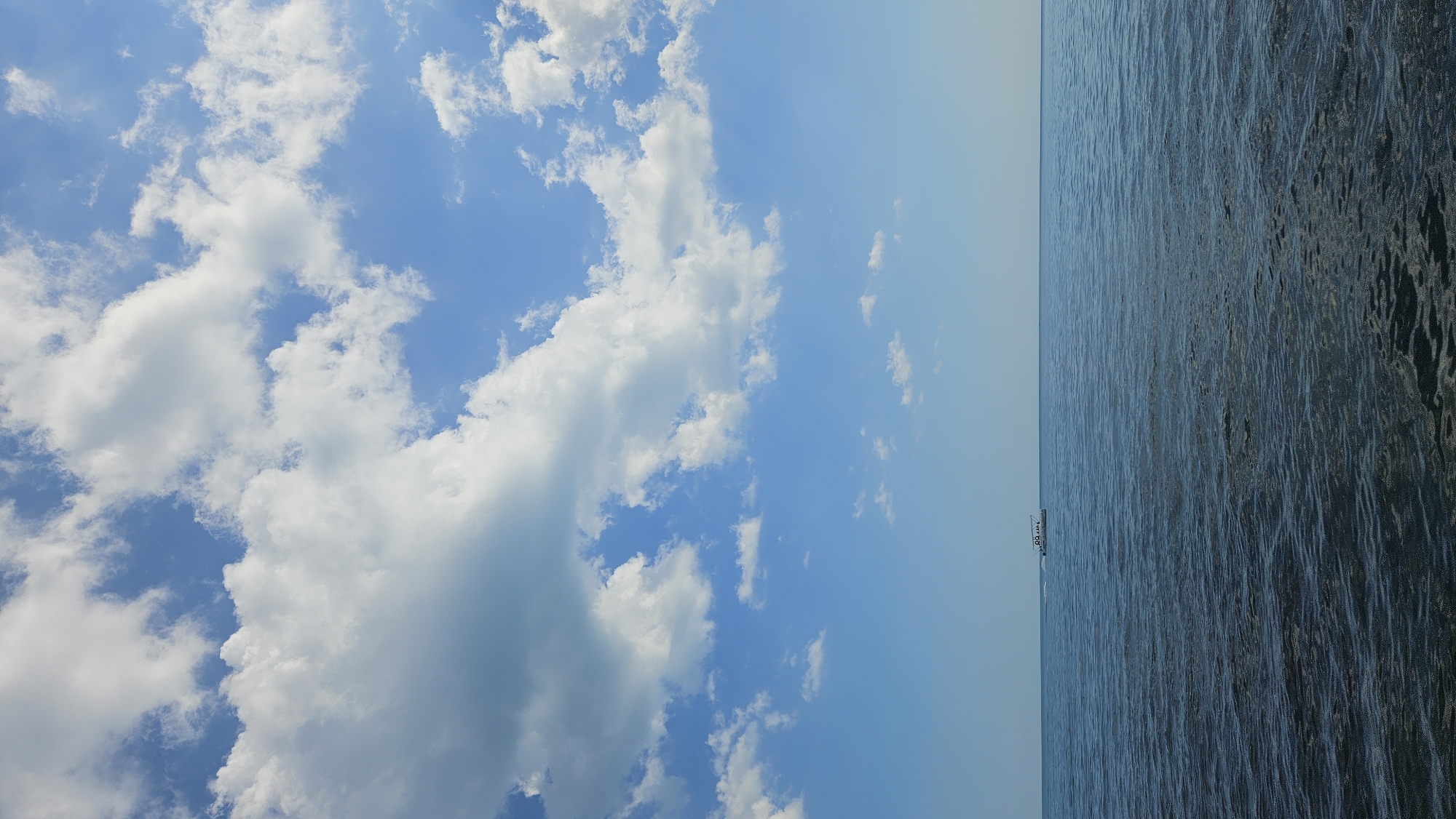 The image depicts a wide shot of the sky and the sea, oriented vertically. The sky, filling the left half of the frame, is a vibrant blue dotted with fluffy white clouds of various sizes. The clouds are scattered, creating a dynamic and airy atmosphere.
On the right side of the image, the deep blue sea stretches to the horizon, with subtle ripples indicating gentle movement on its surface. The water appears calm and vast. A small white boat with a dark roof is visible in the distance, positioned near the horizon line between the sky and the sea. Its presence suggests a serene day at sea, possibly during daylight hours given the brightness of the sky and the illumination of the clouds.
The overall mood of the image is peaceful and expansive, evoking a sense of tranquility and the natural beauty of a seascape. The time of day appears to be daytime, likely late morning or afternoon, with clear skies and moderate cloud cover. The location is suggested to be near Dyurso, Russia, implying a coastal or maritime setting. No discernible text is visible in the image.