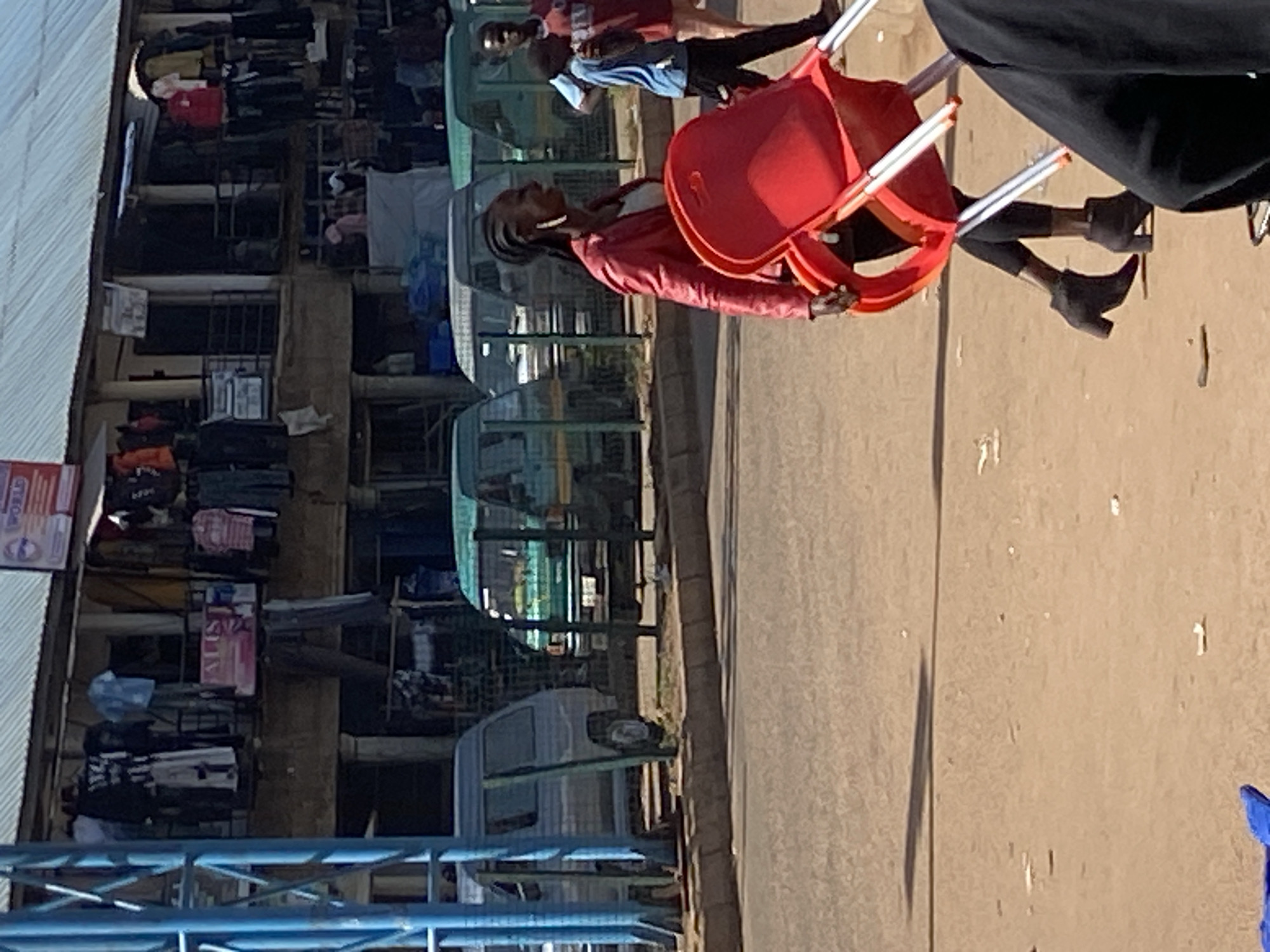 In Bukuru, Nigeria, a woman in a red jacket and dark leggings, wearing ankle boots, walks carrying a red plastic chair. She has braids and is wearing large earrings. Behind her, two men are visible. One man, wearing a red and blue shirt, stands with a child. The other man, wearing a blue shirt and black pants, walks with him. In the background, several vans are parked in front of a marketplace building displaying clothing for sale. A blue metal structure is to the left of the scene. The ground is a dirt road.