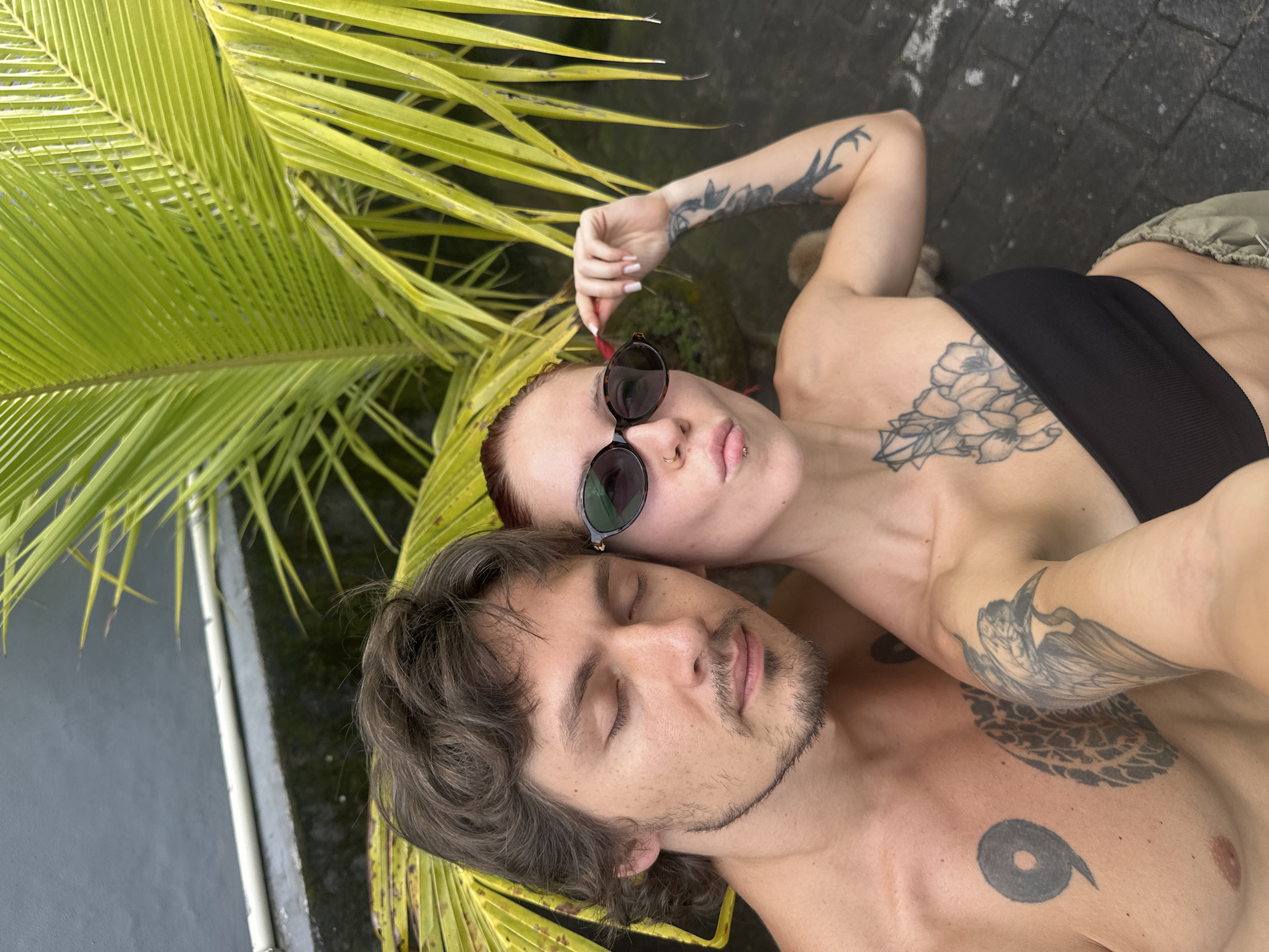 The image depicts two individuals, one male and one female, engaged in a close-up selfie-style photograph outdoors in Dalung, Indonesia.
The male, positioned on the left, has dark brown wavy hair and a scruffy mustache and beard. His eyes are closed. He is bare-chested, displaying a large black "9" tattoo on his left pectoral and a patterned black tattoo below it. A partial tattoo of a bird's head is visible on his left arm.
The female, on the right, has red hair and is wearing dark oval sunglasses. She has a pouting expression with a ring piercing in her left nostril and a labret piercing below her lower lip. Her left hand is raised, adjusting her sunglasses. She wears a black strapless bandeau top and olive green pants. Visible tattoos include a prominent floral design on her chest/sternum and a sleeve tattoo on her left arm depicting branches or antlers. Additional tattoos are partially visible on her right arm.
The background features vibrant green palm fronds or similar tropical foliage. Above the foliage, a dark grey wall or structure is visible with a white horizontal pipe or rail. The ground below the individuals appears to be a dark, possibly wet, paved surface. A small, light-brown animal is partially obscured behind the female's right shoulder.