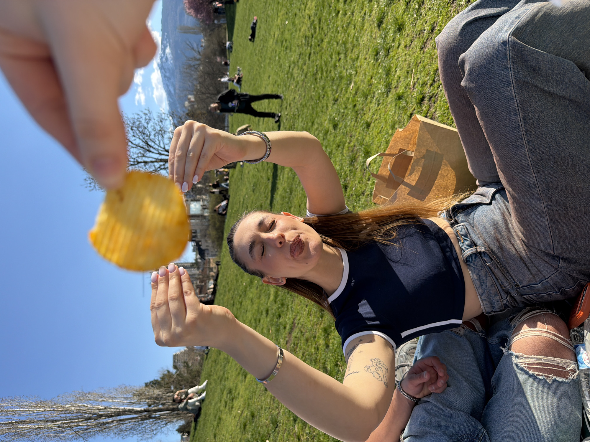 A wide shot shows a young woman with long, brown hair seated on the grass in Borisova gradina, a park in Sofia, Bulgaria. She wears a dark blue crop top with white trim and distressed denim jeans. A man, seated beside her, is also wearing blue jeans. The woman is positioned to eat a potato chip, held by a hand out of the frame. Several other people are scattered across the green grass in the background. A brown paper bag and water bottle rest next to the couple on the grass. In the distance, there are mountains and blue sky.