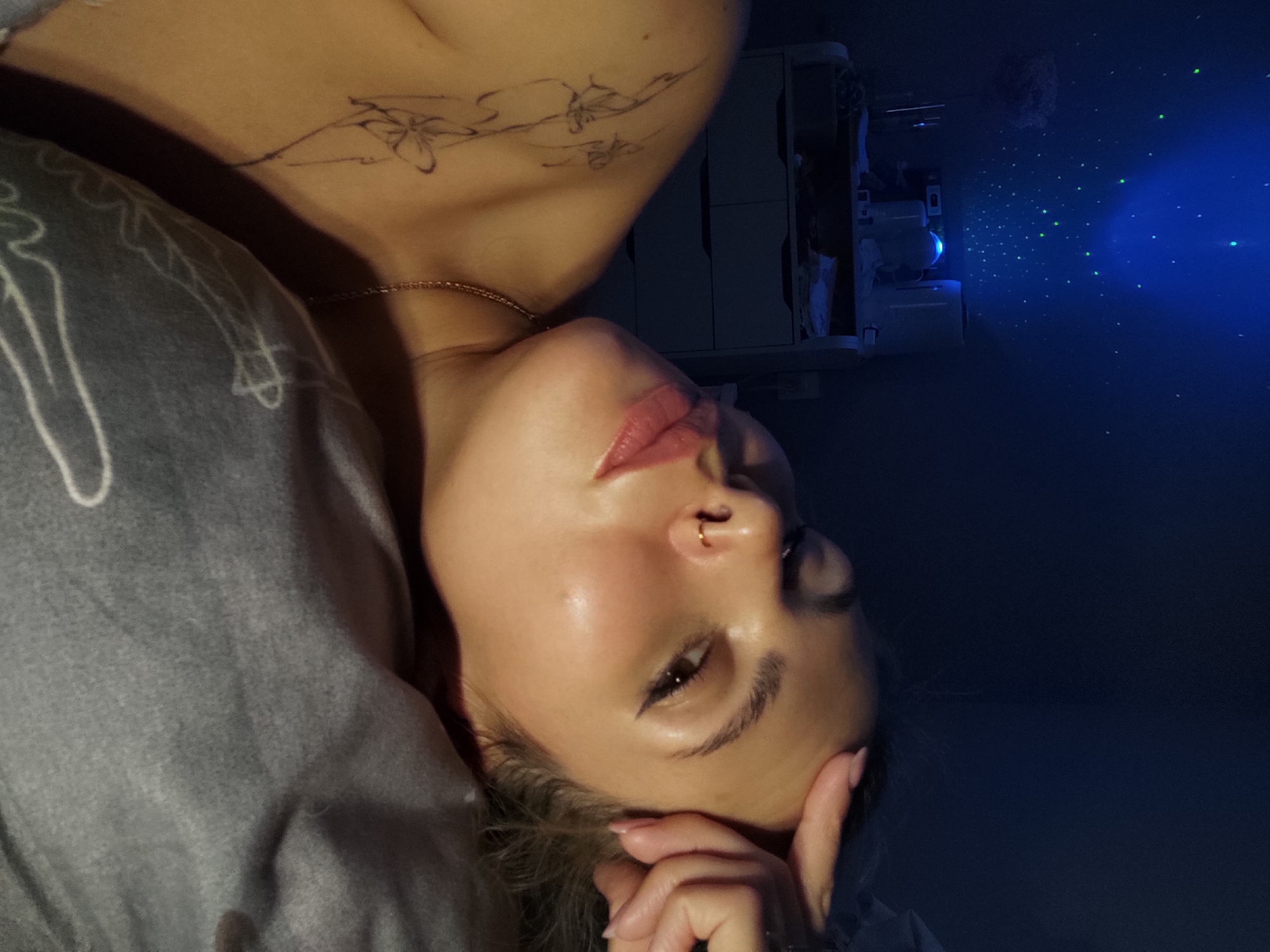 A young woman is lying down in what appears to be a dimly lit room. She is facing upwards with her head resting on her hand. She has a nose ring and a light gold necklace. A tattoo is visible on her chest, below her neck. There is a projection screen in the background displaying blue and green lights. It is impossible to tell if the location is Chaikovsky, Russia.
