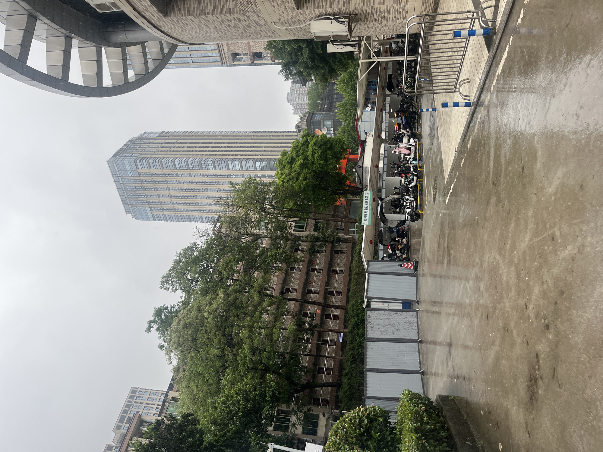 This vertical image captures an urban scene in Yuzhong District, China, on an overcast or rainy day, as evidenced by the wet, reflective paved surfaces.
A prominent, modern skyscraper with a glass facade dominates the background, rising high into the grey sky. To its right, an older-looking building is partially visible through a canopy of lush green trees that punctuate the urban landscape. In the mid-ground, a paved area, likely a street or courtyard, stretches towards the foreground.
On the right side of the frame, a designated parking area houses numerous scooters and motorbikes beneath a protective shelter. A metal barrier, featuring blue and white striped poles, separates this parking zone from the main paved area. A green sign attached to the barrier clearly displays Chinese characters: "校园电动自行车充电区", which translates to "Campus electric bicycle charging area", indicating a campus setting.
On the far left, part of a building with a distinctive curved, metallic architectural element is visible. Further down, other urban structures, including one with a brick-textured wall featuring mounted air conditioning units, are present. Low hedges and utilitarian metal barriers line parts of the foreground, adding to the urban, possibly construction-adjacent, atmosphere. The sky is uniformly grey, suggesting a cloudy day without direct sunlight.