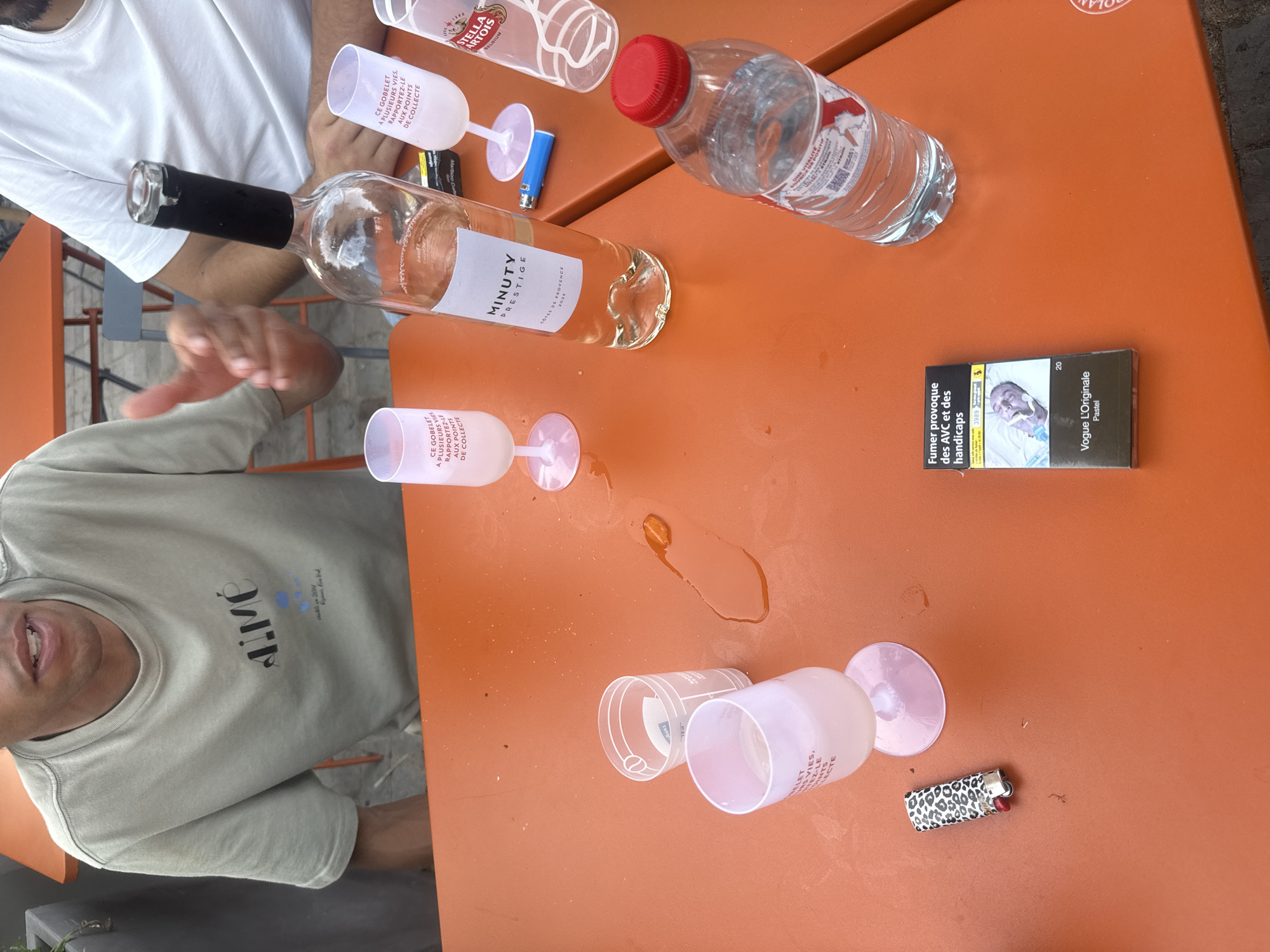 The image shows a table in an outdoor setting. Two people are partially visible. One is wearing a white t-shirt and another is wearing a light green t-shirt. On the table are a bottle of Minuty Prestige rosé wine, a bottle of water with a red cap, two Stella Artois branded plastic cups, and three more generic plastic wine glasses, some of which are empty and one spilled. There are two lighters (one is blue and one has an animal print), and a pack of Vogue L'Originale Pastel cigarettes. The text printed on the wine glasses mentions "CE GOBELET A PLUSIEURS VIES" and "AUX POINTS DE COLLECTE," indicating they are reusable and should be returned to collection points. The cigarette pack has a warning about strokes and disabilities. The table is orange. The location is most likely Paris, France because the cigarette packaging uses the French warning and the beer brand is commonly available.