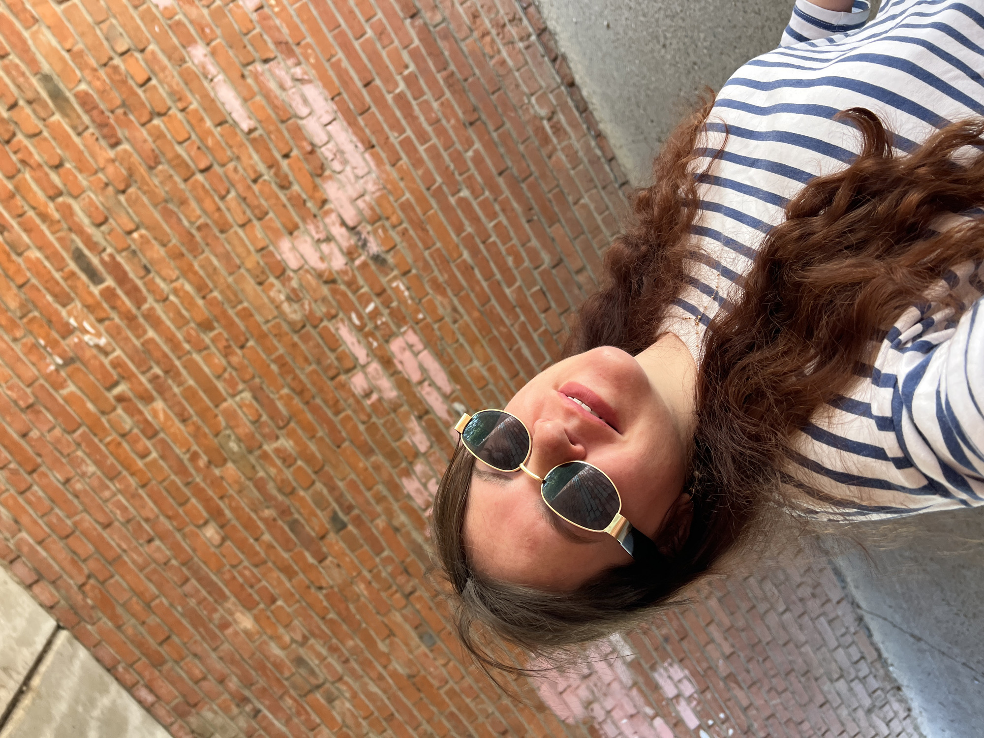 A woman wearing sunglasses and a striped shirt leans her head back against a brick wall. She is outdoors, with the brick wall forming the background of the image. The lighting suggests it is daytime. The image does not provide enough context to determine the location within Noviy, Russia.