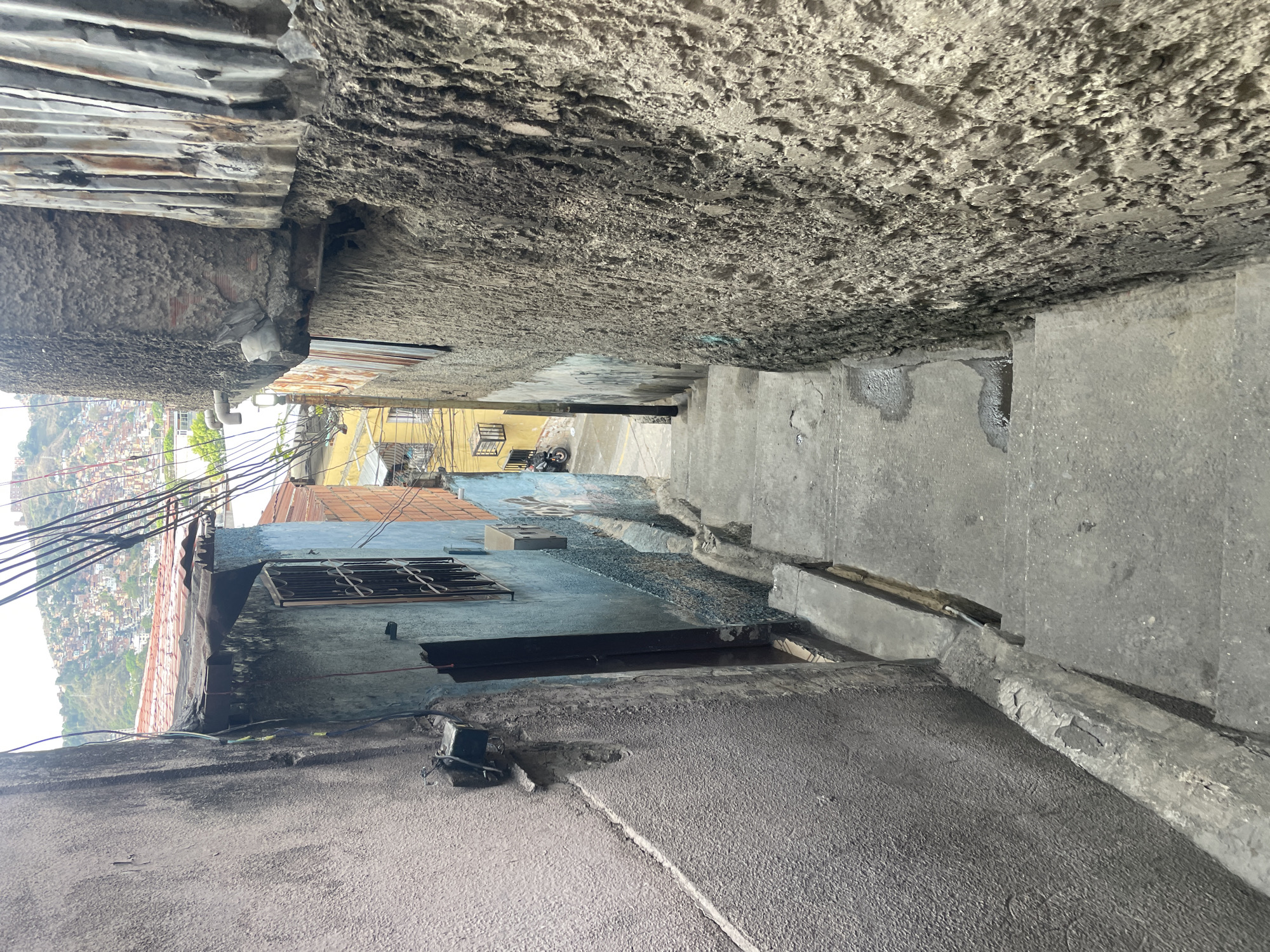 This image depicts a narrow passageway or alleyway in Caracas, Venezuela, characterized by steep inclines and rough, textured concrete surfaces. Above, there is a mixture of corrugated metal sheets and exposed concrete forming the ceiling. Below, there is a descending series of steps and sloped surfaces that comprise the passage. Further down the alley, a few buildings are visible. On the left side of the image, there is a glimpse of the cityscape and the sky beyond the alley entrance. Electrical wires are also visible, traversing the upper left of the image. A barred window on a blue building is visible on the left. The overall condition of the environment suggests urban decay or a low-income neighborhood. No people are visible.