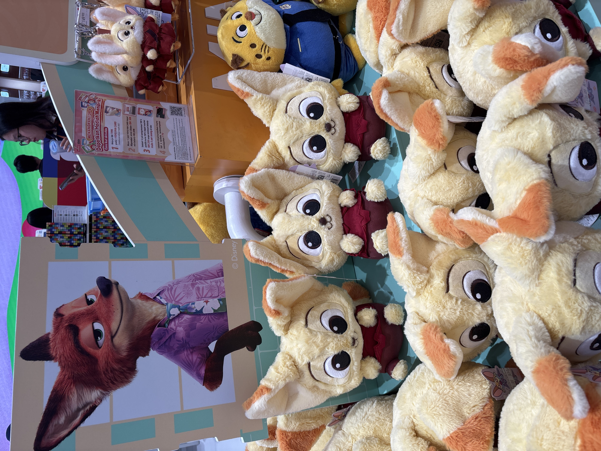 The image depicts a display of plush toys, likely in a retail setting in West Jakarta, Indonesia. In the background, a large cutout of Nick Wilde from Disney's Zootopia is visible, wearing a pink floral shirt and tie. In the mid-ground, several identical plush toys resembling fennec foxes with large eyes and orange-tipped ears are arranged, some wearing red vests. To the right of these, a plush toy of a character resembling a tiger wearing a blue police uniform is also displayed. In the foreground, a pile of similar fennec fox plush toys are seen, with their heads and ears visible. A promotional sign with Indonesian text is visible to the left of the rabbit plush toys, and a price tag is hanging from one of them. A woman wearing glasses and holding a phone is partially visible in the upper right quadrant of the image, observing the display. The overall environment appears to be an indoor store with brightly lit displays.