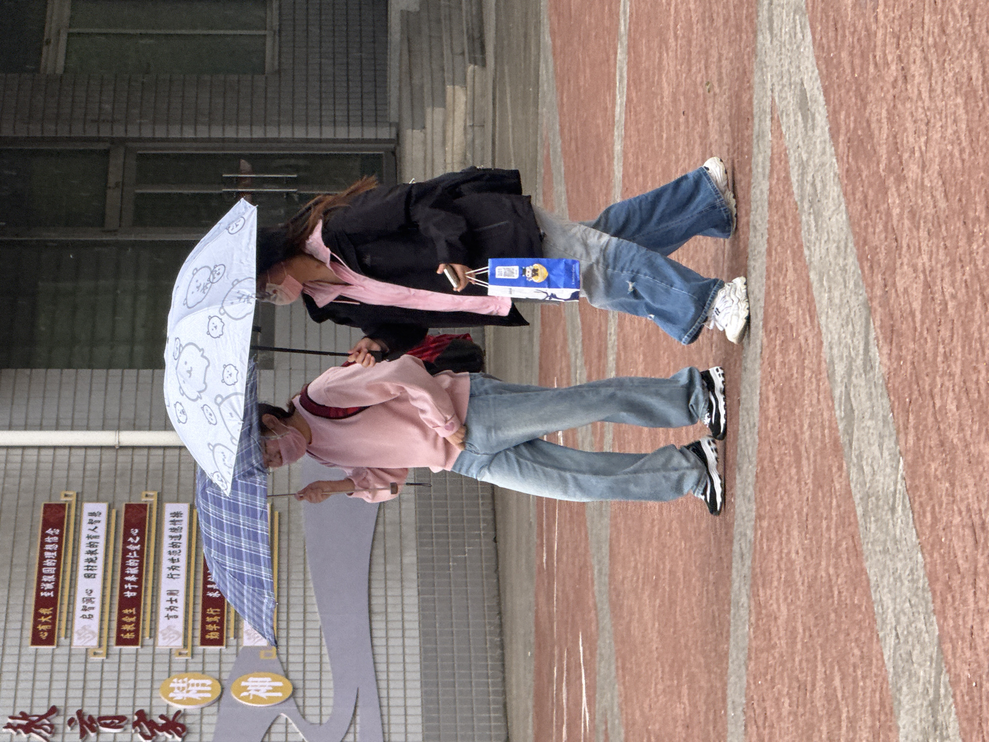 Two young women are walking side-by-side on a red-bricked sidewalk in front of a school building in Zhongtun, China. Both are wearing face masks and carrying umbrellas. The woman on the left wears a pink long-sleeved shirt, jeans, and black-and-white sneakers. She holds a partially open blue and white plaid umbrella. The woman on the right wears a black jacket over a pink shirt, jeans, and white sneakers. She holds a blue and yellow package. The school building behind them has a brick facade and a sign in Chinese characters.