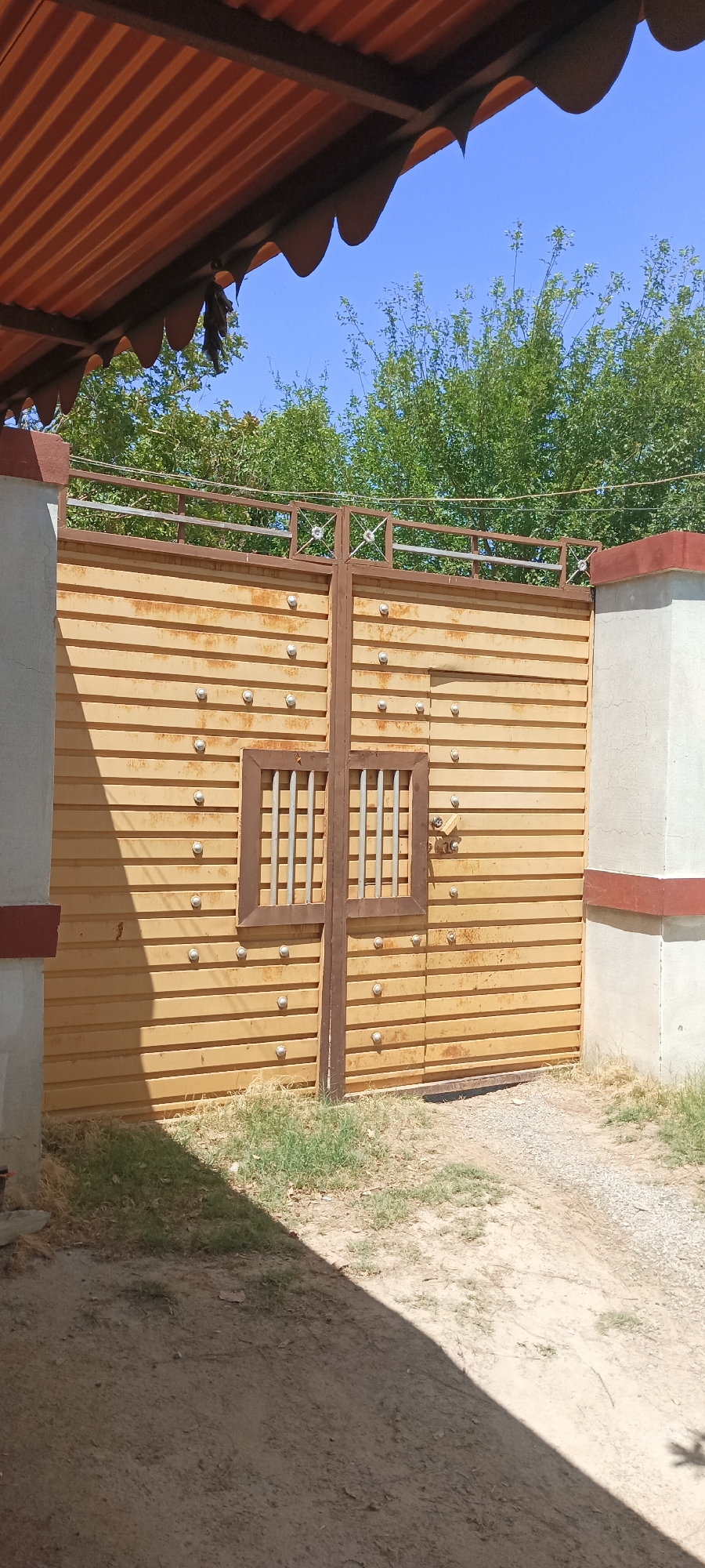 The image depicts a horizontal metal gate in Katlang Tehsil, Pakistan. The gate is composed of closely spaced horizontal slats with a small, barred window in the center and vertical supports. Rust is visible on the gate's surface. The gate is flanked by two white pillars, each trimmed with a red stripe at the top and bottom. The ground in front of the gate is a mix of dirt, gravel, and sparse grass. A partially visible roof structure is present in the upper left of the frame. Trees and a clear blue sky are visible above the gate.