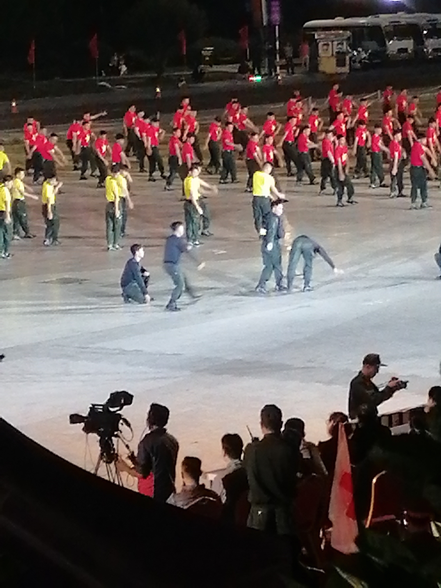 The media file depicts an outdoor evening event, possibly a performance or ceremony, taking place in Da Lat, Vietnam. A large group of individuals, mostly men dressed in dark pants and red or yellow shirts, are engaged in synchronized movements, suggesting a choreographed routine. Some are standing, while others are in various stages of motion, with arms extended or bent. In the foreground, a cameraman operates a large video camera mounted on a tripod. Several other people are visible in the foreground, some observing the event and others involved in its production or management. The scene is illuminated by artificial lighting, creating a nighttime atmosphere. The background shows a blurred expanse, possibly a large open area or plaza, with hints of structures and vehicles, suggesting an urban or event venue setting. The overall mood appears to be one of organized activity and public gathering. There is no visible text in the image.