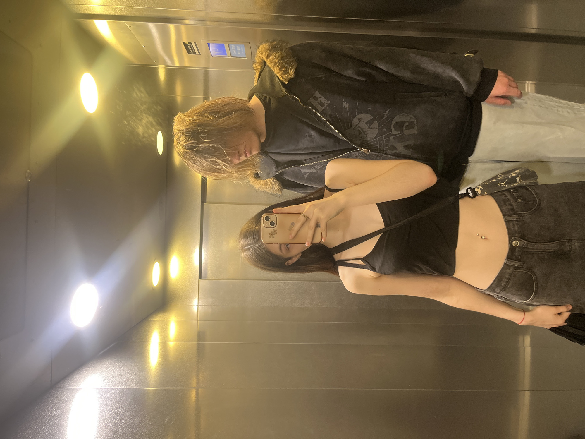Two young adults are captured in a mirror selfie within an elevator in Saint Petersburg, Russia. The female on the left, with long dark hair, holds a gold-colored smartphone with a floral-patterned case, obscuring her face. She wears a black spaghetti strap crop top, dark grey jeans, a red string bracelet on her left wrist, and displays a belly button piercing. A small, patterned black bag hangs from her left shoulder. To her right, a male with long, shaggy light brown hair, which covers most of his face, stands looking downwards and to his left. He is dressed in a dark, fur-lined zip-up hoodie over a dark t-shirt and light blue baggy pants. The elevator interior features reflective brushed metal wall panels and a ceiling with multiple recessed circular lights, some emitting warm yellow light and others bright white light. An elevator control panel with a blue-lit display is visible on the right wall.