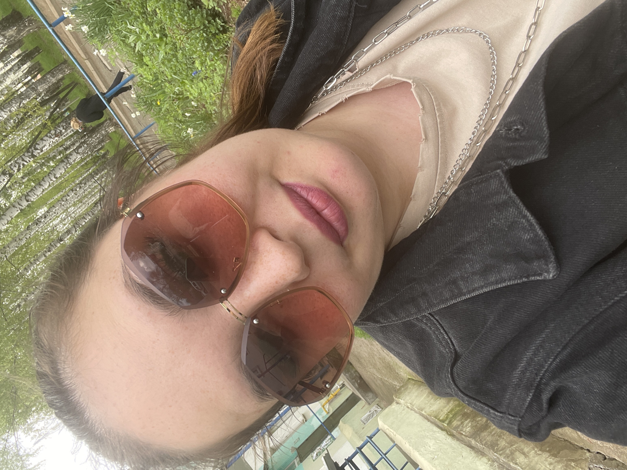 A selfie of a woman wearing sunglasses, a beige t-shirt, and a black denim jacket. She has a silver chain necklace. The background shows trees and a body of water. Based on visual clues such as tree types and architectural details, the location is Chaikovsky, Russia. A person in black attire is visible in the background, possibly walking.