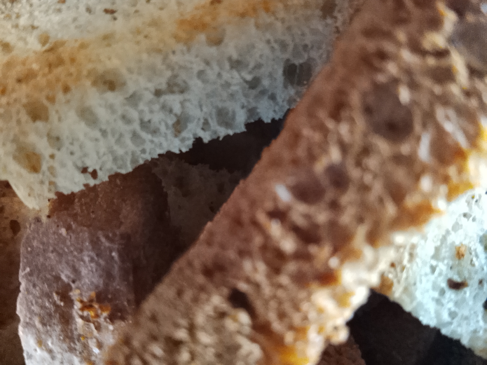 The image is a close-up, macro shot of sliced bread. The bread appears to be a rustic loaf, with visible air pockets and a crust that varies in color from light brown to darker, toasted hues. There are multiple slices overlapping each other, creating a layered composition. The texture of the bread is a primary focus, with its porous crumb and slightly uneven surface. The lighting is soft, highlighting the details of the bread's interior and exterior. There are no people or any elements that would indicate a specific location within the city of Almaty, Kazakhstan. The image solely depicts slices of bread.