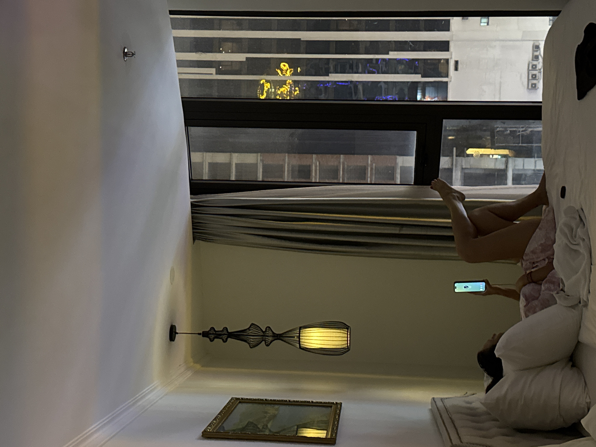 A woman is lying in bed in a room in Đà Nẵng, Vietnam, using a smartphone. The room has a large window with a view of city buildings. A picture frame is on the wall below a light fixture.