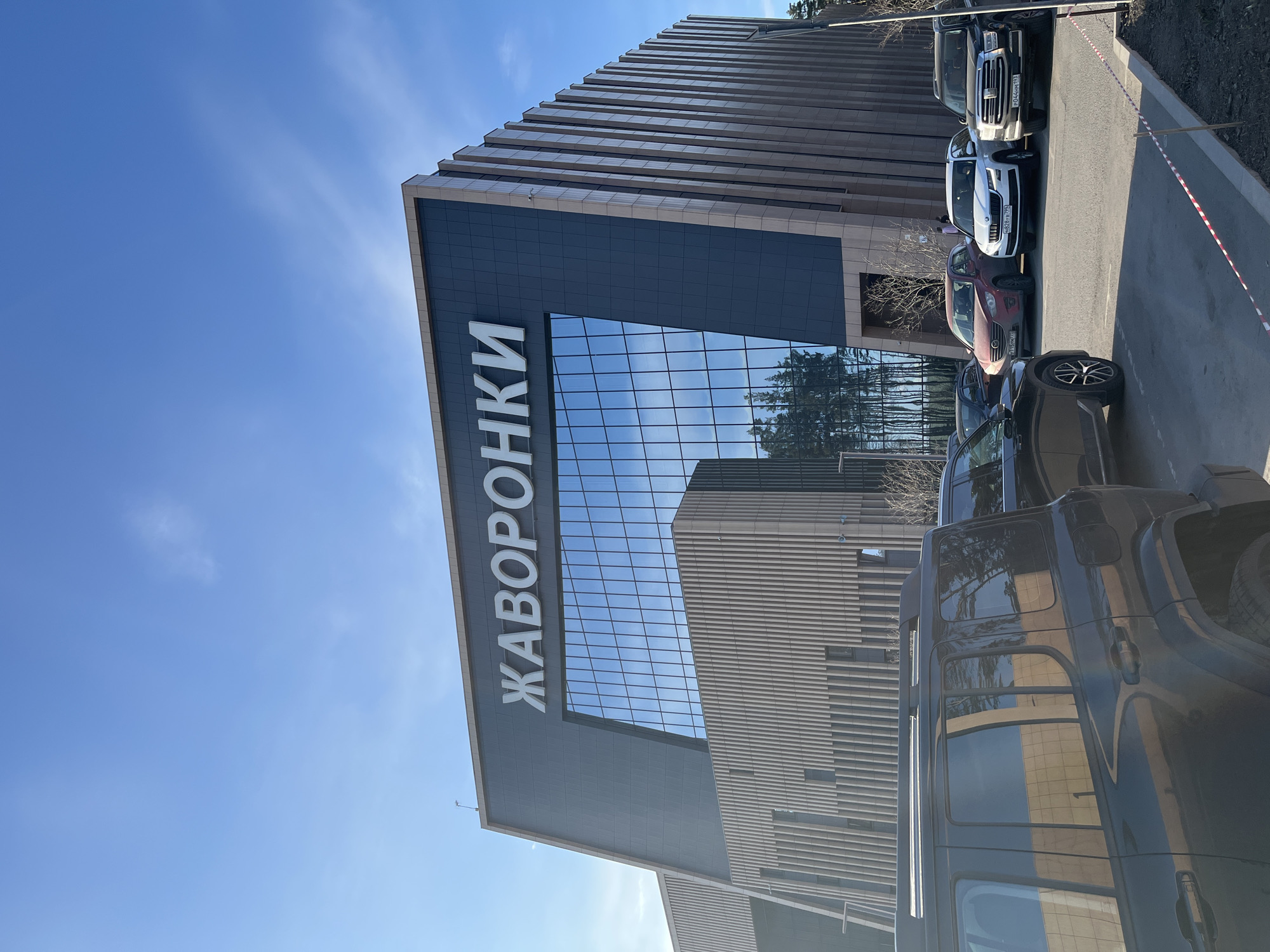 A low-angle shot depicts a multi-story building with a prominent sign in Cyrillic, "ЖАВОРОНКИ" (Zhavoronki), against a clear blue sky. The building's facade features alternating horizontal bands of light and dark materials, with a large glass section reflecting the sky and trees. A row of parked cars lines the right side of the frame, varying in color and model. A strip of red and white barrier tape runs along the edge of the parking area. The building is located in Zhavoronki, Russia.