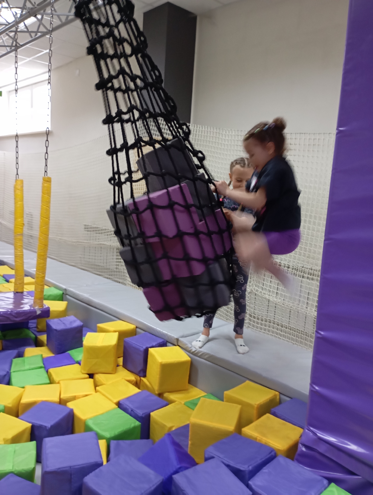 The image depicts a scene at what appears to be an indoor trampoline park. Two young girls are interacting with a suspended net structure filled with gray and purple foam blocks. One girl is in mid-air, seemingly jumping from or towards the net. She is wearing a black shirt and purple leggings. The other girl is standing on the edge of the trampoline area, near the net, wearing a patterned outfit and white shoes. Below the net structure, there is a pit filled with yellow, green, and purple foam cubes. A chain with a yellow protective cover is visible in the background. The trampoline area is bordered by a white mesh net. A purple padded structure is located on the right of the picture. A gray column is in the background.