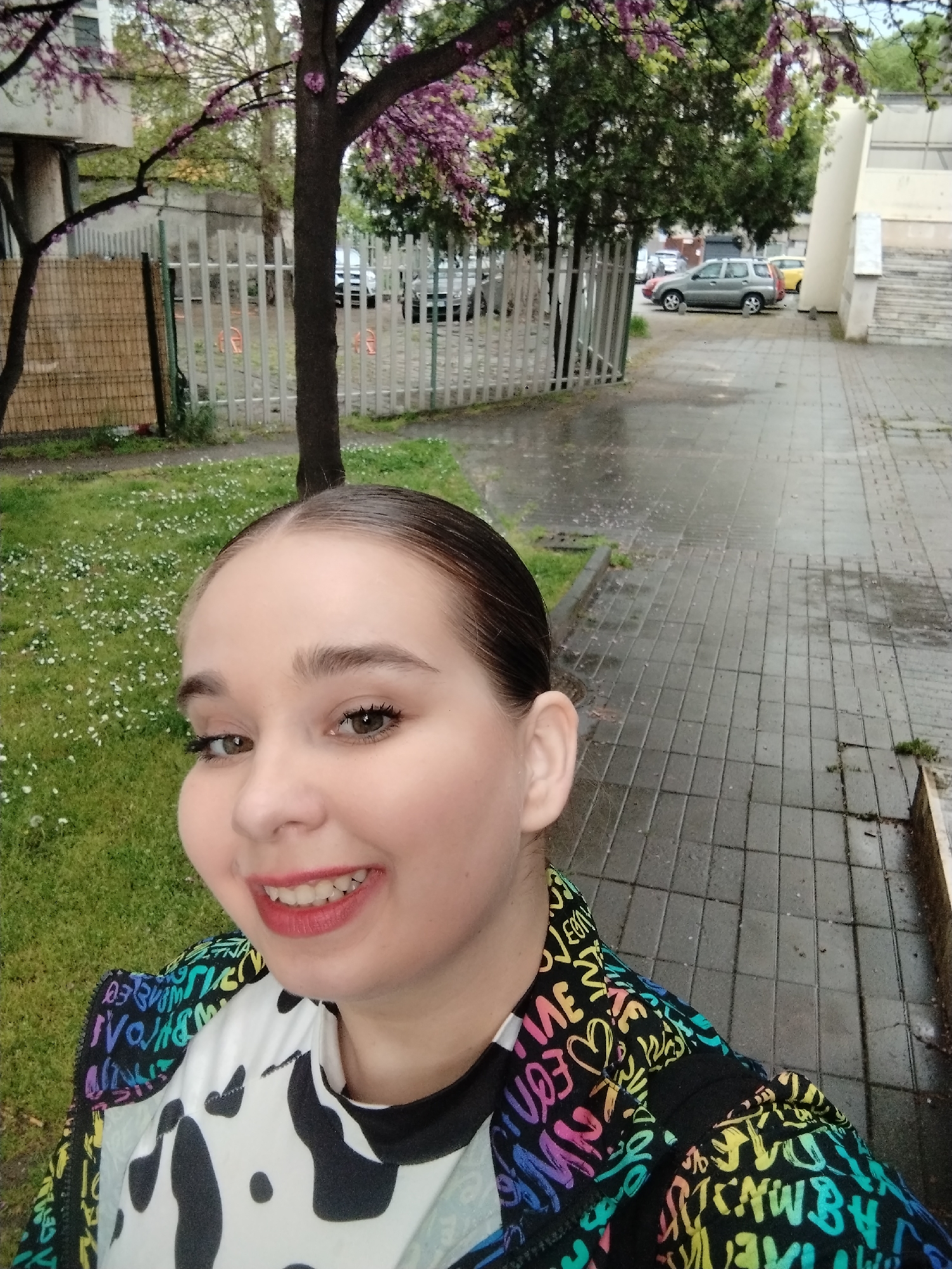 A close-up selfie captures a young female smiling brightly, positioned in the foreground of an outdoor scene in Plovdiv, Bulgaria. She has light skin, dark hair pulled back, and wears makeup, including dark eyeliner and red lipstick. She is dressed in a white top with a black cow-print pattern, layered under a vibrant jacket adorned with multi-colored, graffiti-style text. Visible words on her jacket include "LOVE," "ART," "HAPPY," and "LIFE," among others, in various fonts and a rainbow palette.
The background suggests an urban or park-like environment. To the left, there's a patch of green grass interspersed with small white flowers, bordered by a fence partially made of woven wood and partially of white vertical metal bars. A prominent tree with distinctive pink and purple blossoms, likely a Redbud or Judas tree, stands in the mid-ground. The pavement in the scene appears wet, indicating recent rain or drizzle. On the right, a dark-tiled sidewalk extends into the distance, where several parked cars, including a silver SUV and a yellow vehicle, are visible beyond a white fence. Parts of buildings and more trees are scattered throughout the background. The lighting is soft and diffused, characteristic of an overcast day.