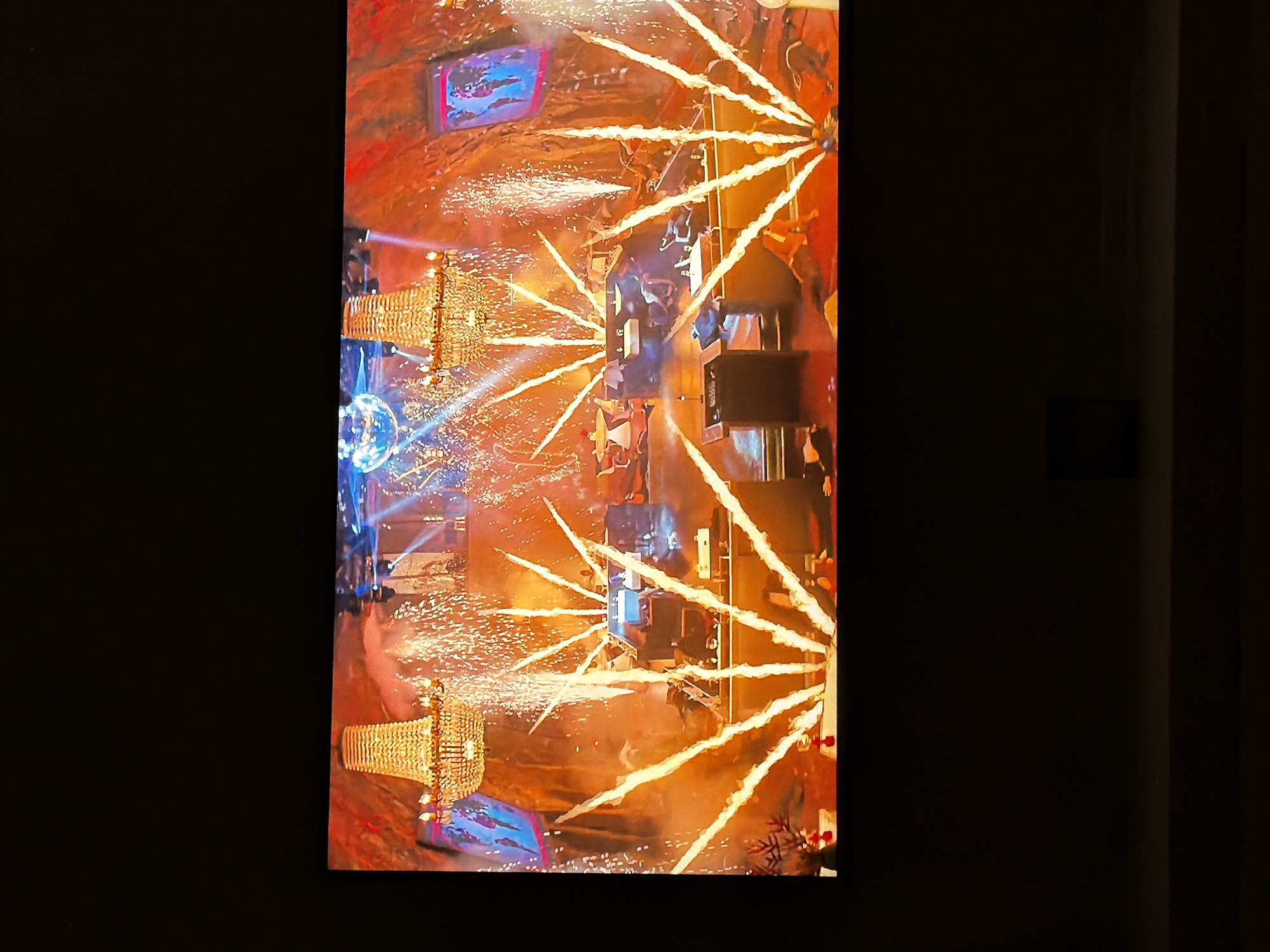 This vibrant image captures an energetic indoor event, likely a celebration or stage performance, in Sofiivska Borshchahivka, Ukraine. The scene is dominated by brilliant special effects and warm, golden lighting.
In the center, multiple streams of golden sparks erupt upwards and outwards from various points on what appears to be a stage or platform, creating a dynamic, starburst-like effect. These pyrotechnics or sparklers illuminate the area with bright orange and yellow light, and numerous small light specks fill the air.
Two ornate, golden chandeliers, appearing large and elaborate, are positioned on either side of the central activity, adding a sense of grandeur. One is visible on the left side, and another is partially visible on the right. Near the left chandelier, a disco ball reflects blue light, contributing to the festive atmosphere. In the background, two large rectangular screens display images with shades of blue and purple, possibly showing abstract visuals or event graphics.
Several people are visible in the central area, amidst the sparks and light. One individual, possibly a performer or host, stands out wearing a white hat or headdress and holding an object resembling a megaphone or trophy. Other figures are less distinct due to the bright light and motion. Dark, rectangular stage equipment, potentially speakers, are also visible.
The overall ambiance is lively and celebratory, with intense artificial lighting, primarily warm tones from the special effects contrasting with some cool blue accents. No discernible text is visible within the image.