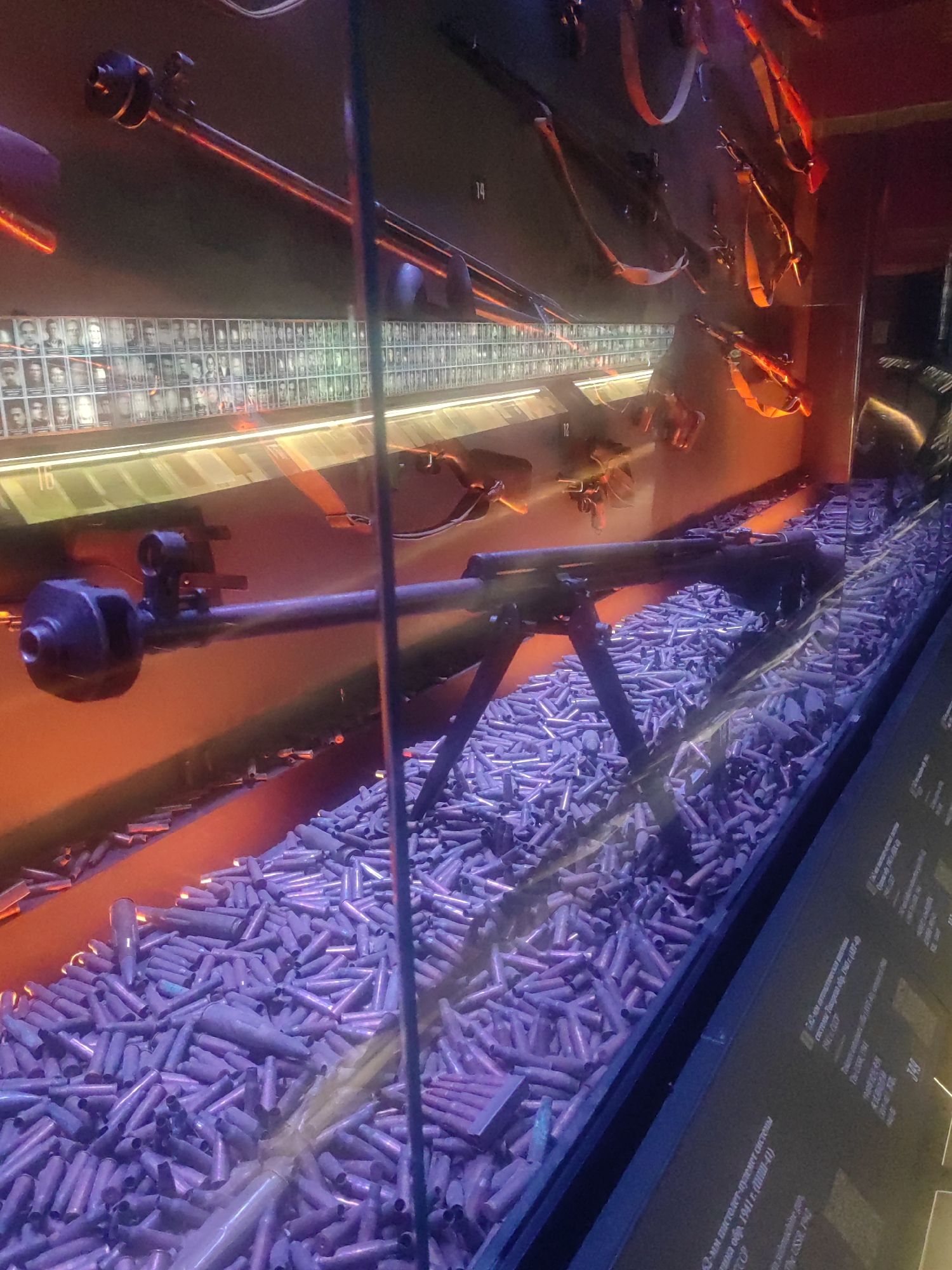 The image depicts a display within a museum setting, likely in Zelenaya Roshcha, Russia. The primary focus is a large machine gun mounted on a bipod, positioned in the foreground and angled upwards. It rests on a substantial pile of spent bullet casings, which fill the lower portion of the display case. The casings appear to be of a uniform size and color, suggesting they are all from the same caliber of ammunition.
Behind the machine gun, a wall is lined with numerous framed photographs of individuals, likely portraits of soldiers or historical figures. These photographs are arranged in a grid pattern and are illuminated, casting a glow on the wall. Above this display of photographs, several firearms, possibly rifles or submachine guns, are hung on the wall by straps, some of which are red.
The overall lighting of the display is dramatic, with warm orange and red hues dominating the background and a cool purple light illuminating the bullet casings. This lighting scheme creates a stark contrast and emphasizes the historical and potentially somber nature of the exhibit. The presence of the machine gun, bullet casings, and portraits suggests a military or historical theme, possibly related to a conflict or war.