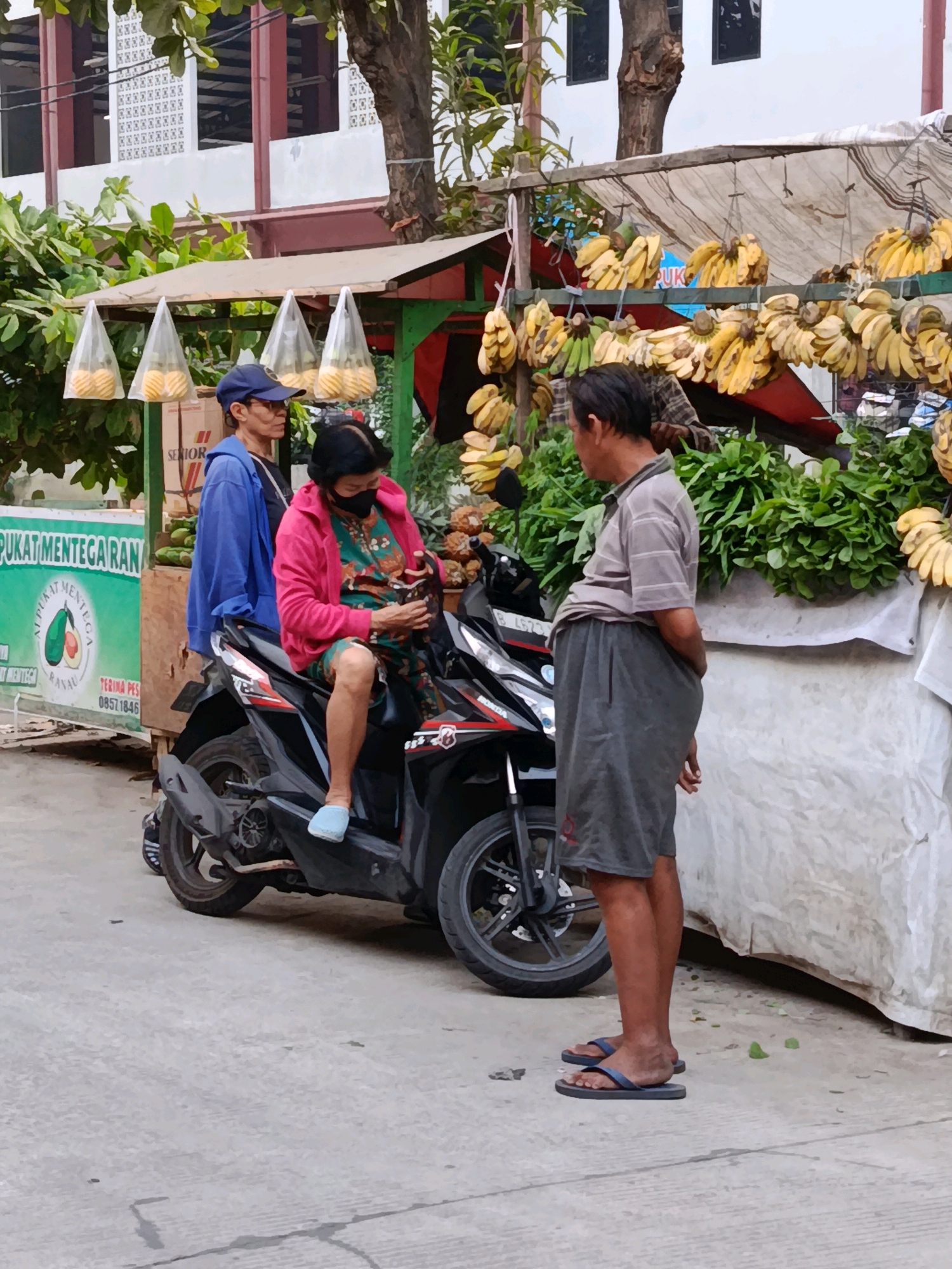 In Bekasi, Indonesia, a street scene captures two individuals by a motorcycle and a fruit stand. A woman, wearing a pink jacket and a green patterned dress, sits on a black motorcycle. She is looking down and holding a dark object, possibly a phone or wallet. Beside her, a man in a blue hoodie and a blue cap stands. To the right, a man wearing a grey striped shirt and grey shorts stands facing the woman, with his hands behind his back. Behind the motorcycle and the man in grey is a fruit stand laden with bunches of yellow bananas and leafy green vegetables. The stall is covered by a white tarp. In the background, a building with white and red architectural elements is visible, along with trees. The ground is paved with concrete.
