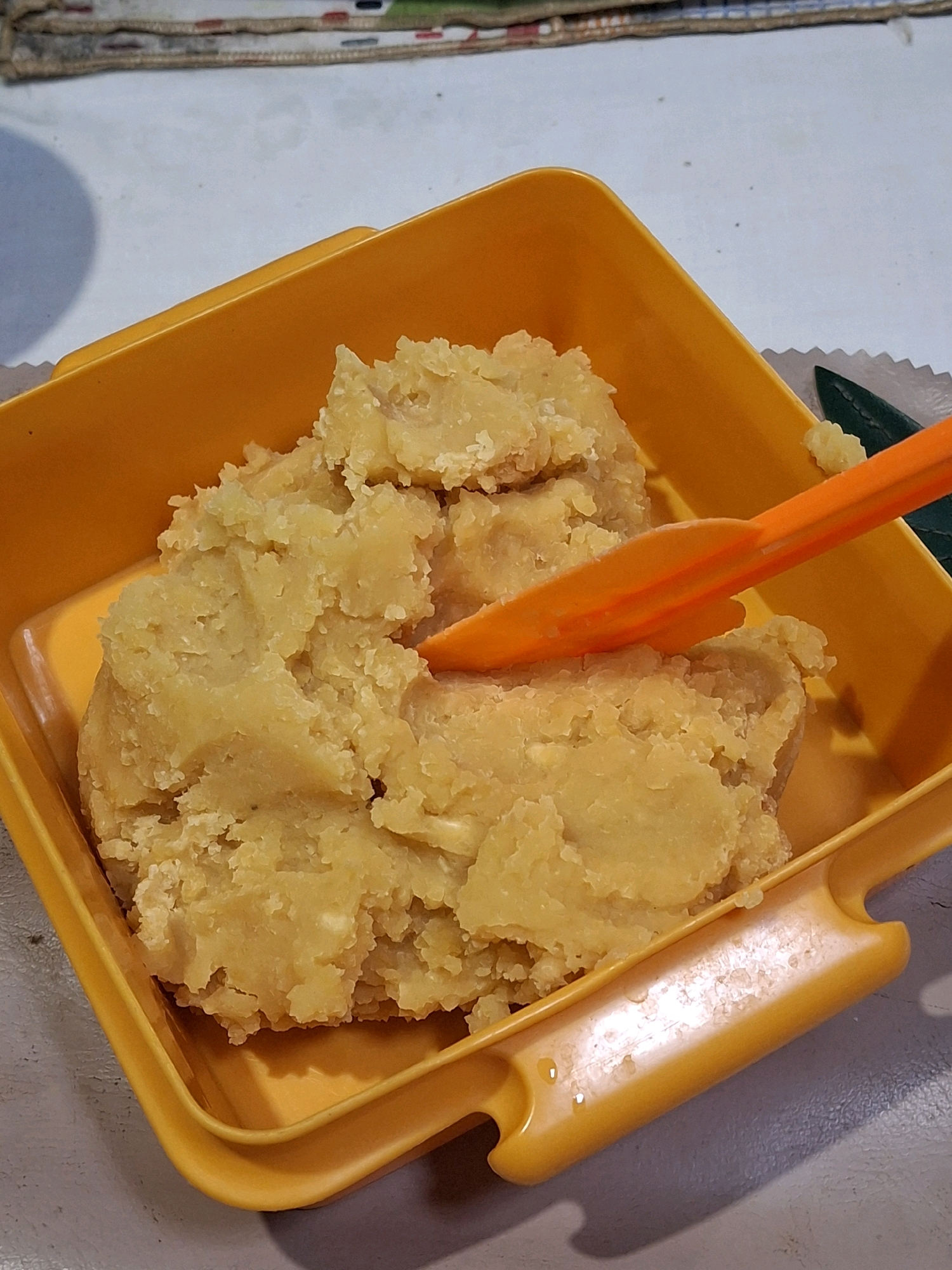 A close-up, high-angle shot shows a yellow plastic container filled with mashed sweet potatoes. An orange plastic utensil is partially submerged in the mashed potatoes, angled towards the right side of the container. The container is positioned on a light gray surface, possibly a table, with a patterned placemat visible in the upper background. The lighting is artificial and casts shadows. There is no indication of the city of Lumajang, Indonesia, within the image.
