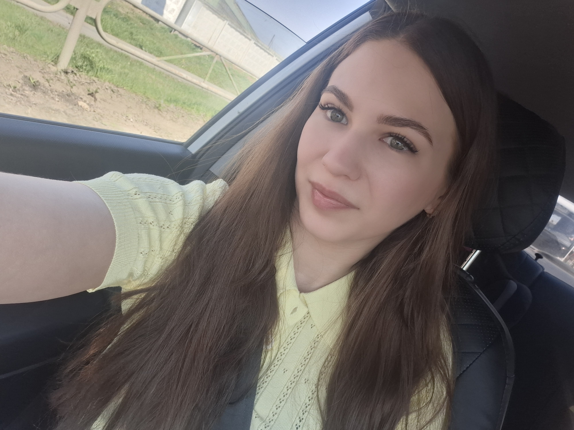 A medium shot shows a woman with long brown hair sitting in the driver's seat of a car. She wears a light yellow collared shirt. The car appears to be moving, as suggested by the blurred background seen through the car window, including greenery, a metal barrier, and a building. The interior of the car is visible, including parts of the steering wheel, dashboard, and passenger seat headrest. The location is potentially in the city of Chaikovsky, Russia, as requested, but this cannot be definitively confirmed from visual information alone.