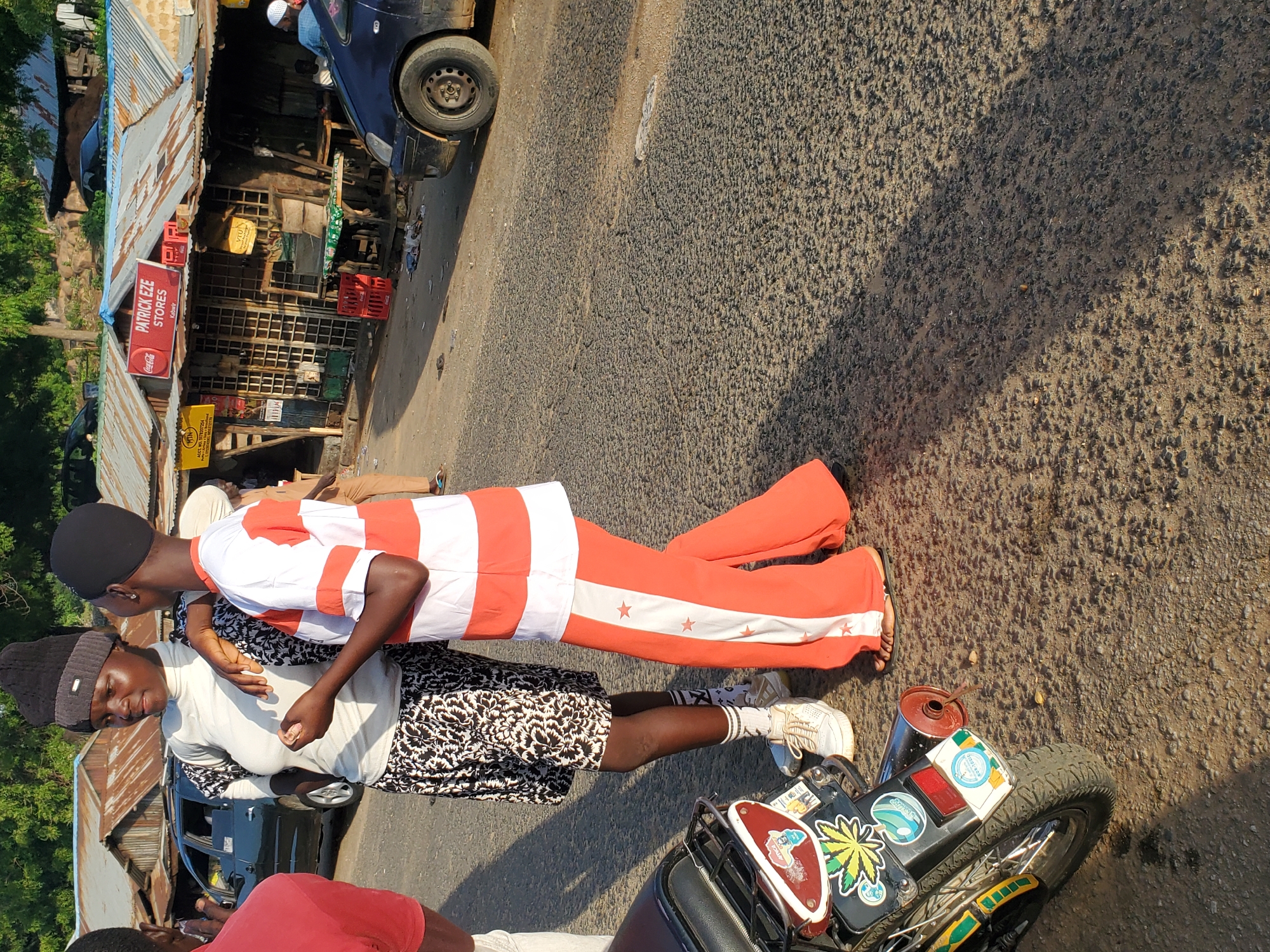 Two young women are standing on a paved road in Ampan, Nigeria, near a row of roadside shops. One woman, on the left, wears a white turtleneck and a black beanie, with a patterned skirt and white sneakers. The other woman, on the right, is dressed in a white and orange striped t-shirt and matching orange pants with white star accents. They appear to be engaged in conversation. Behind them, a dark-colored car is parked, and a motorcycle is partially visible in the foreground on the left. The shops have corrugated metal roofs and display various goods. The overall scene depicts a typical street view in a Nigerian town.
