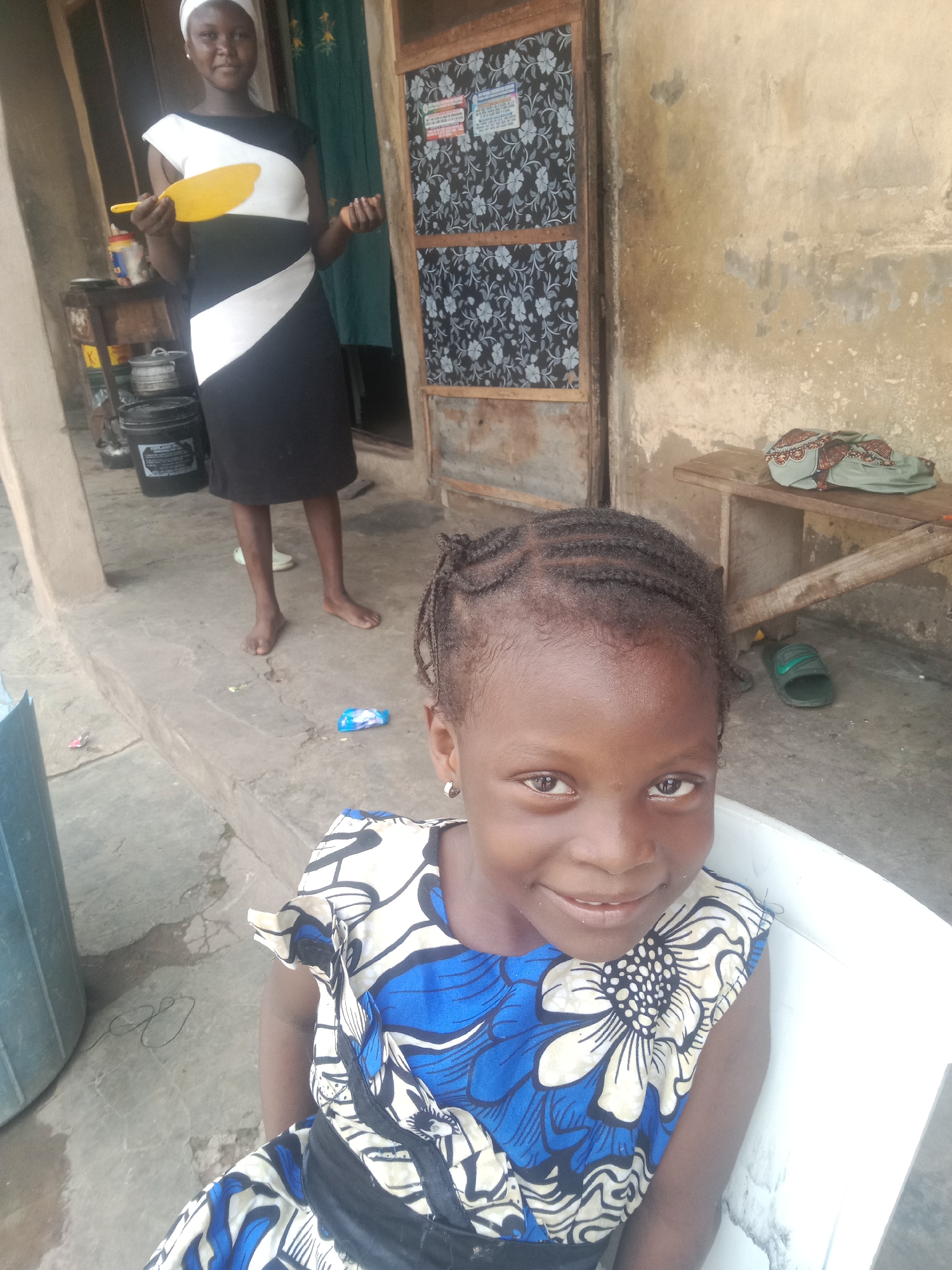 A young girl with dark skin and braided hair is sitting in a white chair, looking at the camera with a slight smile. She is wearing a colorful dress with a blue, white, and black floral pattern. In the background, an older girl or woman wearing a black and white striped dress and a white head covering stands near a doorway. She is holding a yellow object in her left hand and appears to be in the process of doing chores. The environment suggests an outdoor setting, possibly a compound in Lokoja, Nigeria, with a concrete floor, a wooden bench with some fabric draped over it, and a large blue container. The wall behind the subjects is textured and weathered. There are also some objects on the ground, including a green sandal and what appears to be a discarded wrapper. The overall impression is a candid moment captured in a simple, everyday setting.