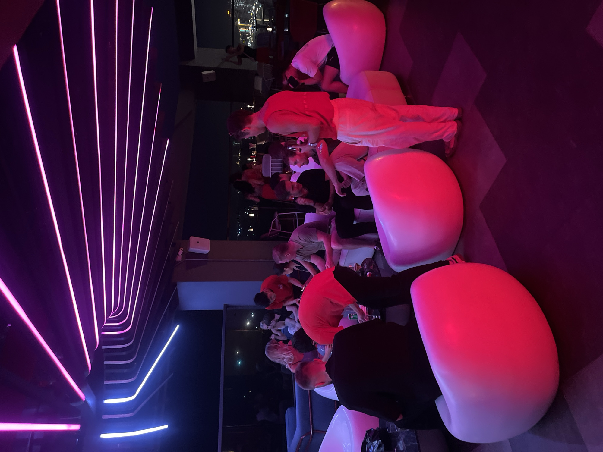 The image depicts a group of people gathered in what appears to be a rooftop bar or lounge with a view of a city at night. The scene is illuminated by vibrant neon pink and red lighting, creating a modern and energetic atmosphere. Several large, egg-shaped chairs or seating units, glowing with pink and red light, are scattered throughout the space.
The individuals are casually dressed and seem to be engaged in conversation or social interaction. Some are seated, while others are standing or leaning. One person in the foreground wears a red sleeveless top and light-colored pants, holding what might be a drink. Another person in a red t-shirt is partially visible. In the background, a person can be seen in what looks like swimwear, perhaps near a pool or a water feature.
The time of day is clearly night, with the city lights of Đà Nẵng visible in the distance, suggesting an urban setting. The weather is not discernible from the image, but the indoor/outdoor nature of the location implies it is likely pleasant enough for an evening gathering. The overall mood seems relaxed and social, typical of a bar or lounge environment. The unique, curved neon lighting installation on the left side of the image is a striking design element. No visible text is present in the image.