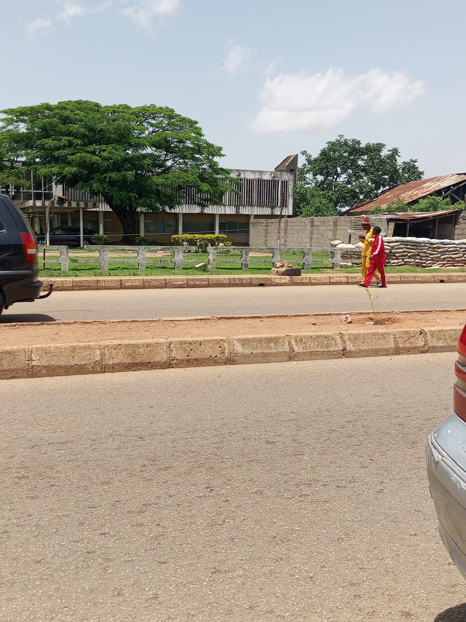 The image depicts a street scene in Bukuru, Nigeria, under a bright, partly cloudy sky. In the foreground, a paved road is visible with a concrete curb and a sandy median. On the median, a discarded plastic cup lies near a small plant. Further back, a dark-colored SUV is parked on the left side of the road. Across the street, two individuals are walking on the sidewalk. One person is dressed in a yellow tracksuit with a red hat, and the other is in a red tracksuit. Behind them, a building with an unusual architectural design featuring vertical concrete elements is situated, partially obscured by a large, leafy green tree. Further to the right, a structure with a rusted corrugated metal roof stands, and sandbags are piled near a brick wall, suggesting a construction or security-related area. The overall atmosphere suggests a warm, possibly arid climate.