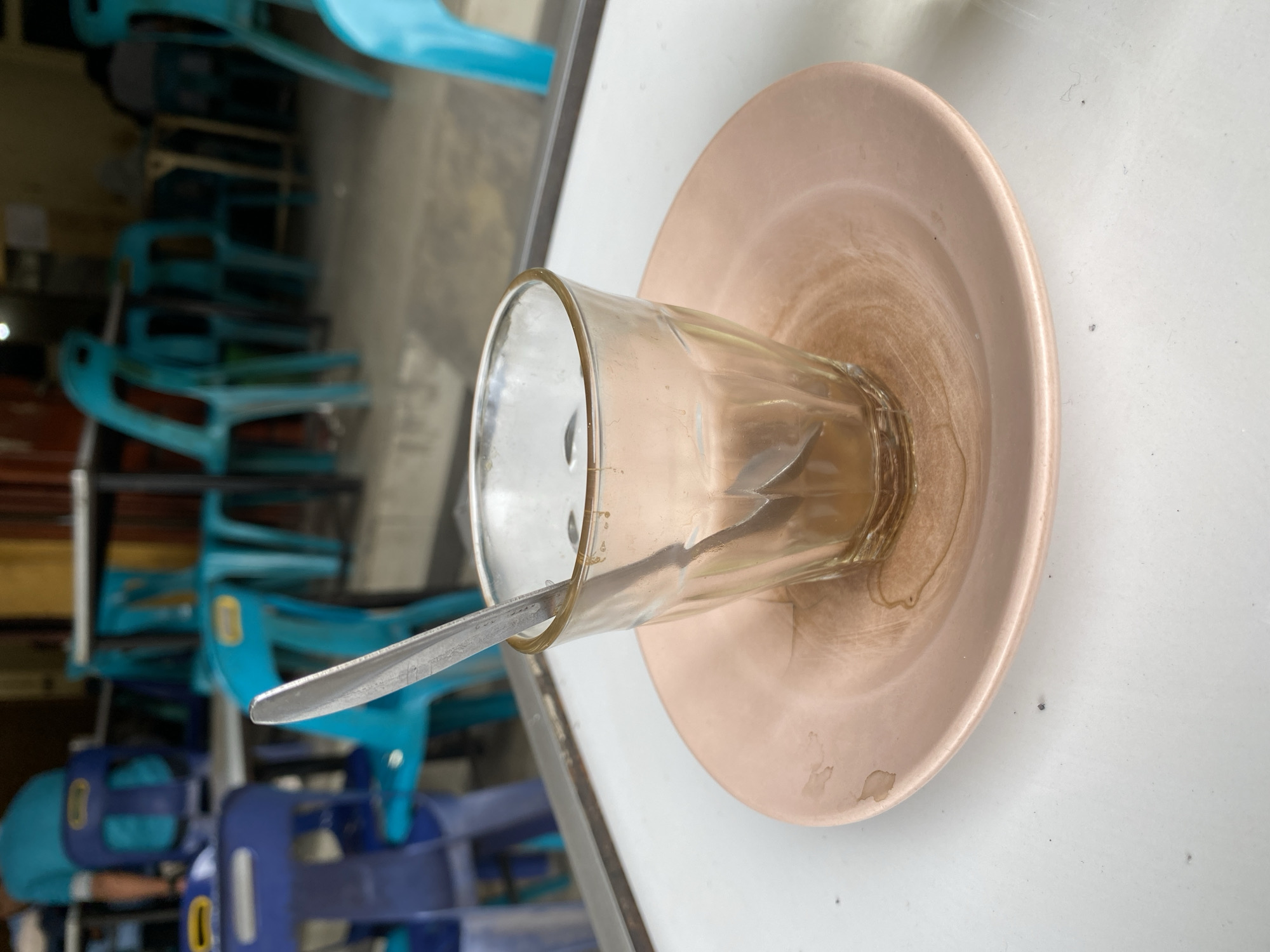 A close-up shot captures an empty, slightly dirty glass of coffee sitting on a pale pink saucer. A spoon rests inside the glass, its handle extending out. The glass has subtle diagonal ridges. The saucer shows evidence of dried coffee stains, forming a circular pattern around the glass's base. The table surface is white. In the blurred background, rows of stacked turquoise and blue plastic chairs are visible, suggesting an outdoor seating area, possibly a cafe or restaurant. The lighting appears to be natural, indicating daytime. The overall impression is a scene of a recently finished beverage at a casual establishment.