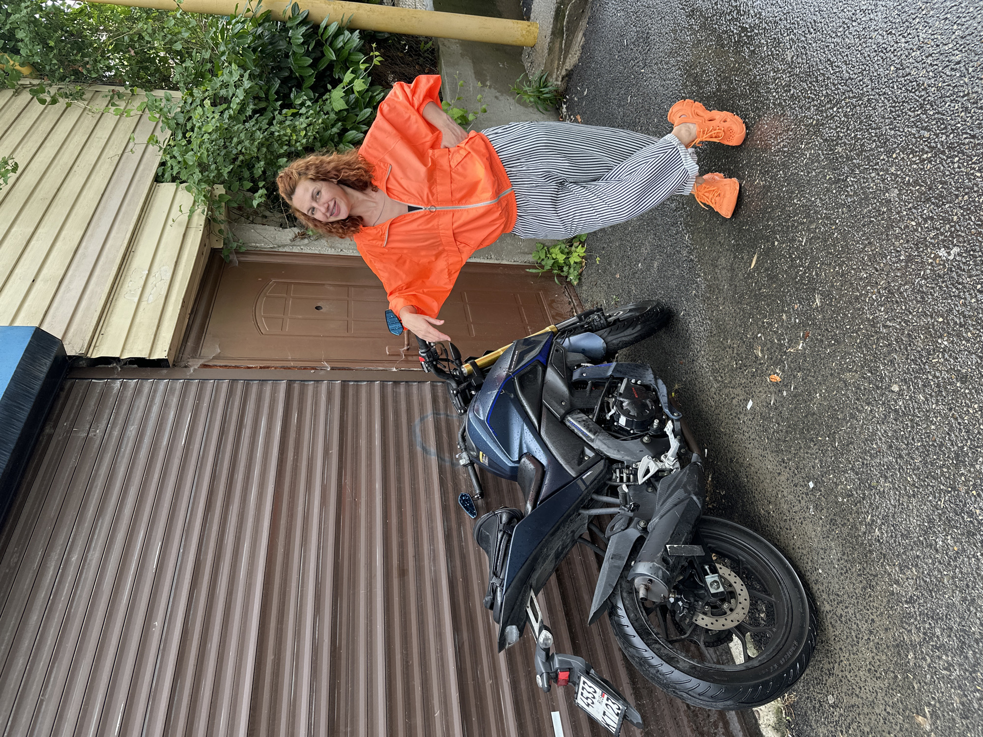 A woman with curly red hair, wearing an orange jacket and black and white striped pants, poses next to a blue motorcycle. The motorcycle is parked on a wet asphalt surface. Behind the woman and motorcycle is a building with a brown corrugated metal garage door and a wooden door. The building is partially covered with green vines. The scene appears to be an urban outdoor setting.