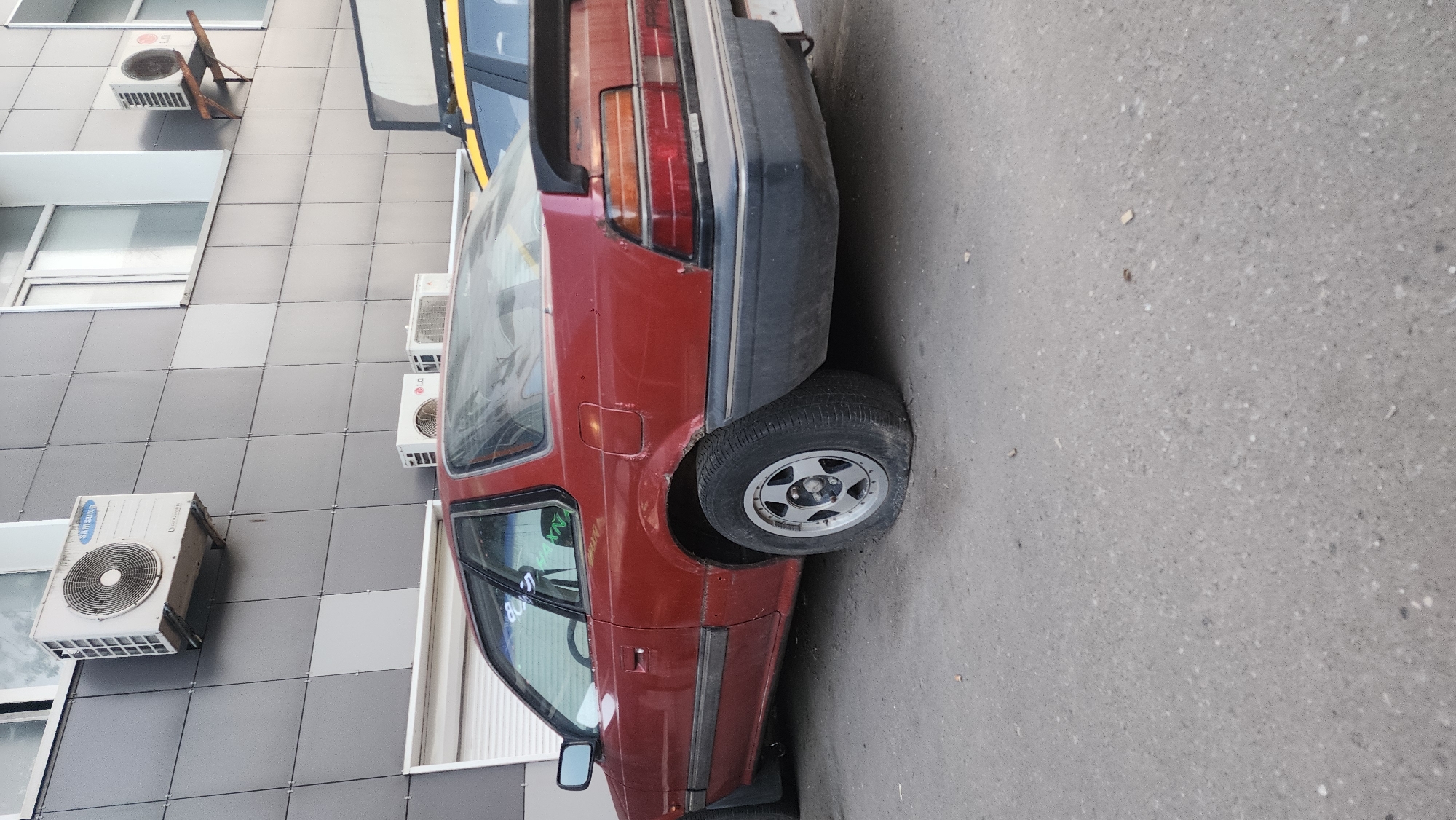 A maroon car with a flat tire is parked on a gray asphalt surface next to a building with a gray and white tiled facade. The building has several air conditioning units attached to its exterior. The location is Moscow, Russia.