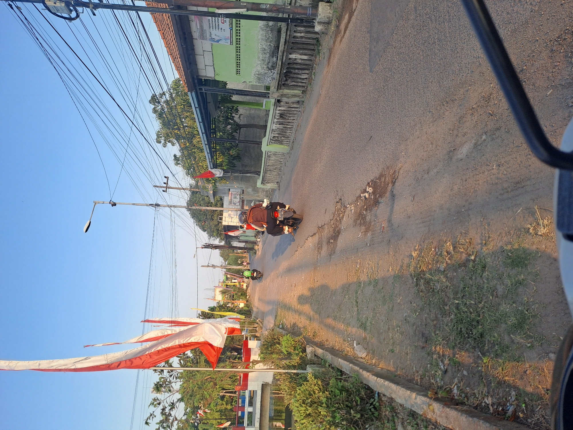 The image depicts a street scene in Pademonegoro, Indonesia, likely during the late afternoon given the long shadows. The perspective is from a vehicle, possibly a motorcycle, looking forward. The road is paved with asphalt and shows signs of wear and tear, with some patches of dirt and debris.
On the left side of the road, there is a drainage ditch or canal with a concrete edge. Lush green bushes and grass grow along the canal's embankment. A white and red bridge with a red railing crosses the canal further down the street. Several Indonesian flags, red and white, are prominently displayed on poles along the left side, suggesting a festive occasion or national celebration. Trees with dense green foliage are visible behind the flags.
On the right side of the road, a row of houses or buildings lines the street. One building is painted light green and has a tiled roof. Another building appears to be under construction or in a state of disrepair, with exposed bricks and a metal awning. Electrical wires are strung overhead, supported by poles, indicating an urban or semi-urban setting. A poster with text and images is affixed to one of the buildings.
Several people on motorcycles are present on the road. Two individuals are clearly visible riding motorcycles in the same direction as the viewer. One motorcycle has a rider wearing a brown jacket and a black helmet, and the other has a rider with a green jacket. The figures cast long shadows on the road due to the low angle of the sun.
The overall environment is a typical Indonesian street scene, with a blend of residential buildings, natural greenery, and evidence of infrastructure. The presence of numerous flags suggests a patriotic atmosphere.