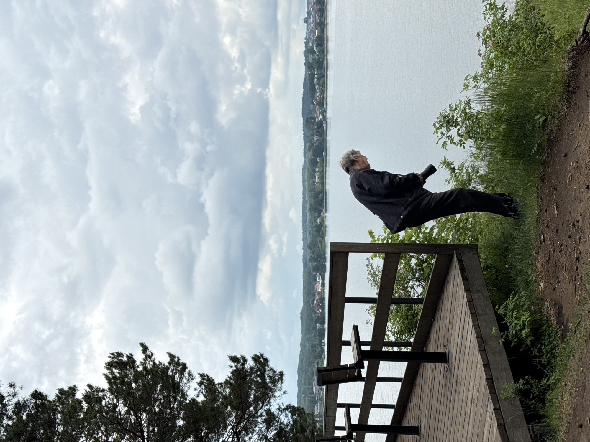 The image captures a scenic overlook of a vast body of water, likely a lake or a wide river, under an overcast sky. In the mid-ground, an individual is positioned on a dirt path, adjacent to a wooden observation deck. This person, with short, light-colored hair and dressed in a dark jacket and pants, is holding a device resembling a camera or binoculars, aimed towards the distant view. They appear to be observing or photographing the landscape.
The wooden platform, featuring prominent railings and a built-in bench, extends towards the water, providing a dedicated viewing area. The immediate foreground features natural elements, including a dirt trail, green grass, and various bushes and trees, some with light pink blossoms. Across the tranquil water, a distant shoreline stretches horizontally, densely covered with green trees and dotted with numerous small houses, indicating a residential or village area. The sky above is largely covered by white and grey clouds, suggesting an overcast day with soft, diffuse lighting. No discernible text is visible within the frame. The scene evokes a quiet moment of appreciation for the expansive natural and semi-urban landscape.