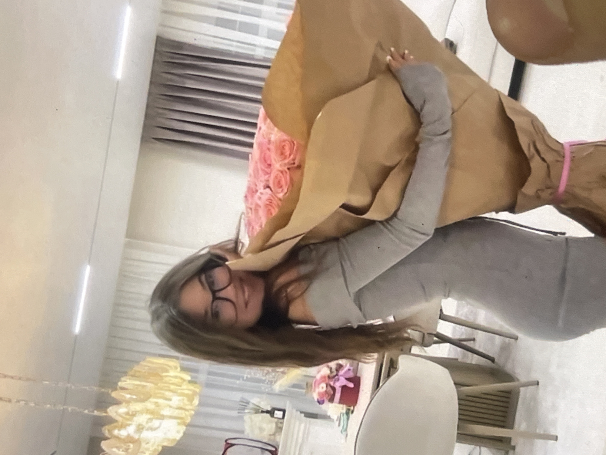 The rotated image captures a young woman holding a large bouquet of pink roses, wrapped in brown kraft paper and secured with a pink ribbon. She is smiling directly at the camera, wearing black-rimmed glasses and a form-fitting, light gray long-sleeved dress. Her long brown hair falls over her shoulders, and her fingernails are painted white or a very pale color.
The scene is set indoors, likely in a modern apartment or home. The background features light-colored walls, horizontal window blinds or shutters, and contemporary furniture including chairs and a table. To the woman's left, a distinctive, layered, light-colored decorative pendant lamp hangs. Further back, on a surface, a smaller floral arrangement in a red box is visible, alongside a diffuser. The bright lighting suggests it could be daytime. The woman appears happy and pleased, possibly having just received the flowers. No explicit text is visible within the image. The location context is Poltava, Ukraine.