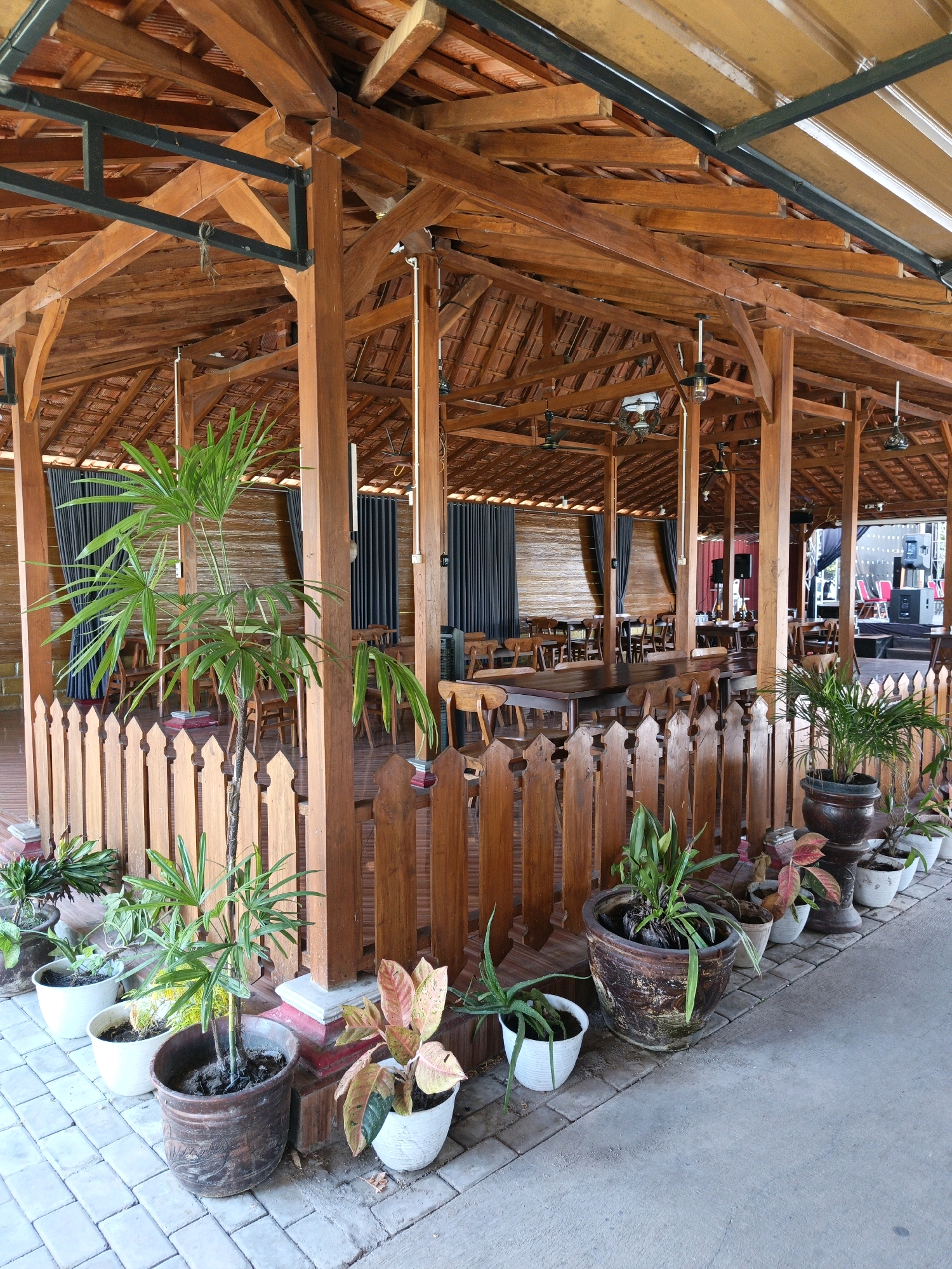 The image shows an outdoor dining area with a wooden structure in Jonggrangan, Indonesia. The structure features a thatched roof supported by thick wooden beams and pillars. A low wooden picket fence separates the dining area from a walkway. Several potted plants are arranged along the walkway and near the fence. In the background, tables and chairs are set up, with dark curtains lining the walls of an adjacent building. The overall environment suggests a casual dining or event space. There are no people visible in the image.