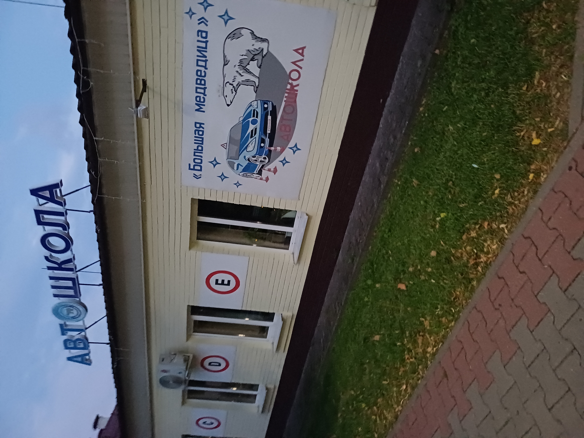The image shows a building with a sign that reads "АВТОШКОЛА" (Autoschool) in large blue letters. Below this sign is a mural depicting a blue car, a polar bear, and cones, with the text "«Большая медведица»" (Big Bear) and "АВТОШКОЛА" (Autoschool) in red. There are circular signs with letters C, D, and E on the building wall next to windows. An air conditioning unit is mounted above a window. The building is light yellow with a dark brown trim at the bottom. A paved walkway with red and gray interlocking bricks runs in front of the building, adjacent to a strip of green grass with fallen leaves. The sky is overcast. The location is the "Big Bear" driving school in Kobryn, Belarus.