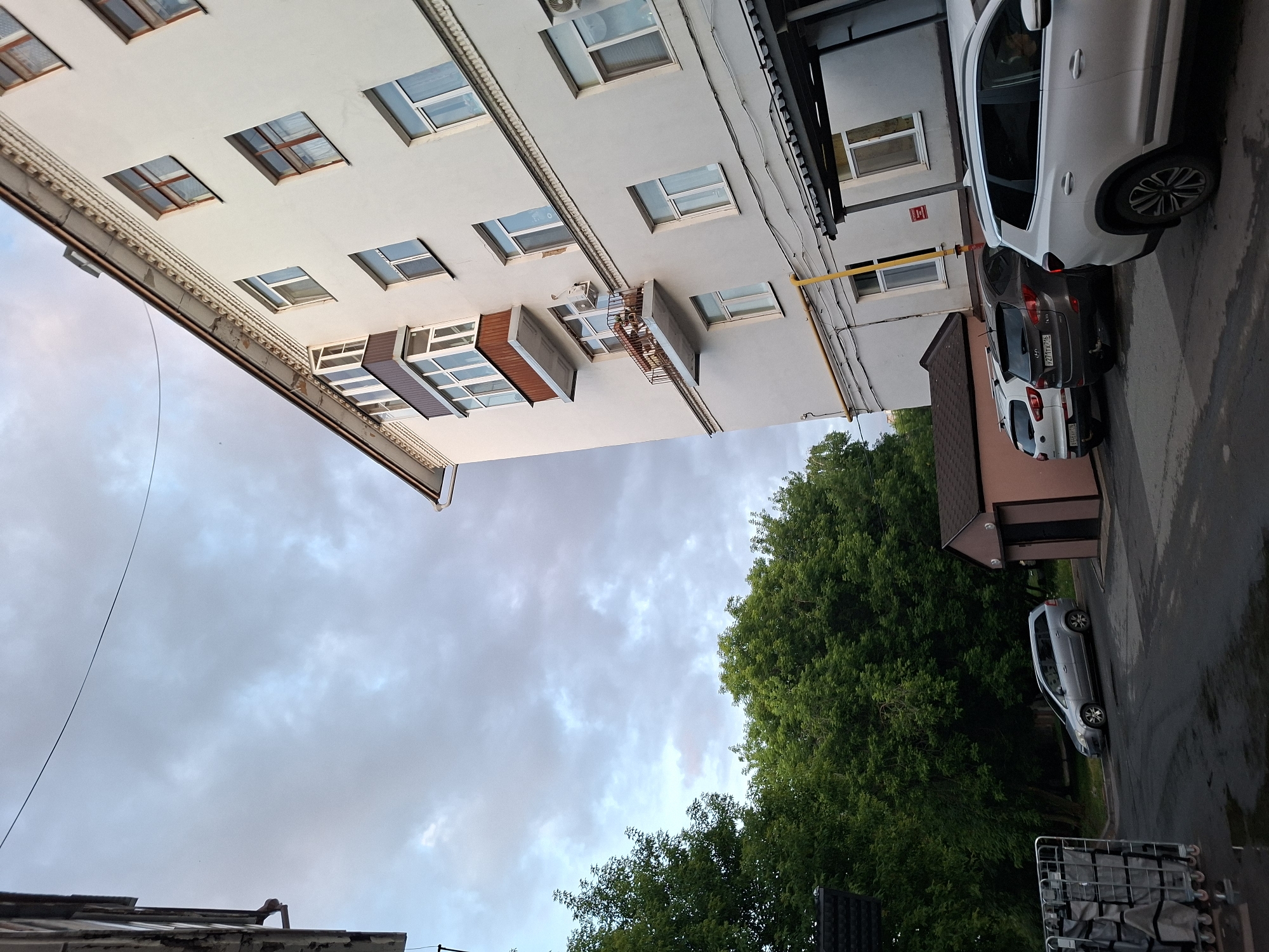 The image shows a view of a white, multi-story apartment building with multiple windows and balconies, viewed from a low angle looking upwards. The building has a decorative cornice near the roofline. A balcony with brown trim and glass panels is visible, as is another smaller balcony with a rusted metal railing and potted plants. The sky above is overcast with gray and light blue clouds.
To the left of the building, there are dense green trees. In front of the building and to the right, several cars are parked: a silver station wagon, a dark grey SUV, a white SUV, and a white crossover. A small, pinkish-brown building with a dark door is situated between the trees and the parked cars. In the foreground, to the far left, a metal rolling cart with grey fabric bags is visible. The ground is paved and appears wet in places. The scene is consistent with a residential courtyard in a city.
The presence of the license plates with Russian characters (e.g., "P277TX 716" and "B466EY 116") indicates the location is in Russia. Based on the architectural style and the license plate format, this scene is consistent with urban residential areas in Kazan, Russia.
